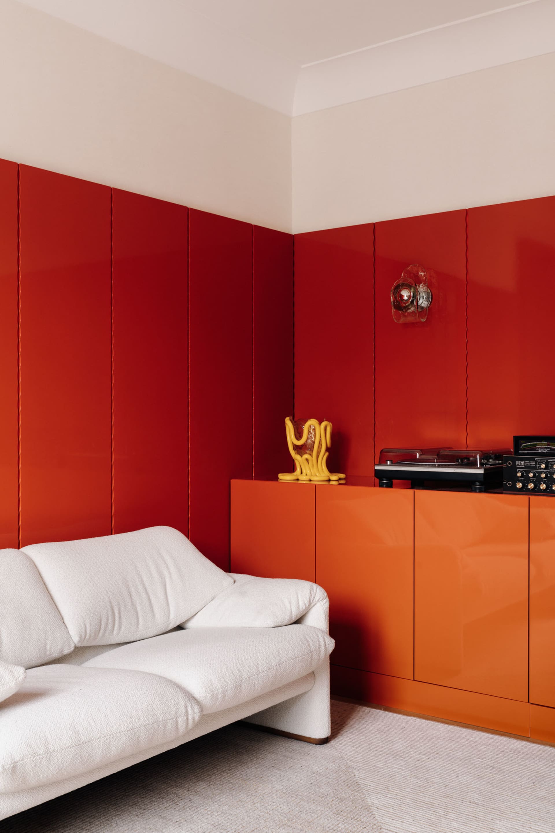 A modern living room with red and orange lacquered paneling, featuring a white sofa and an entertainment center.