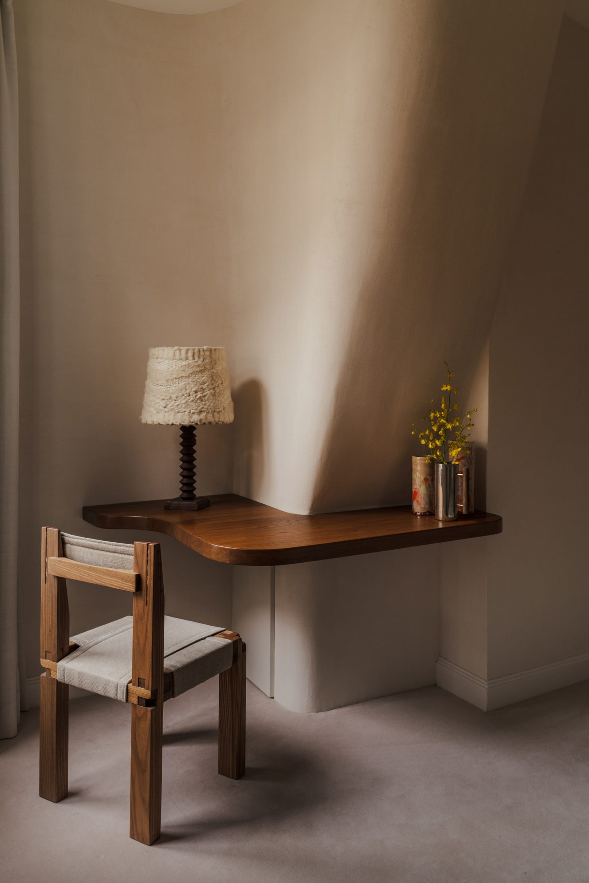 A modern home office setup featuring a wooden desk and chair, with a lamp and flowers on the desk.