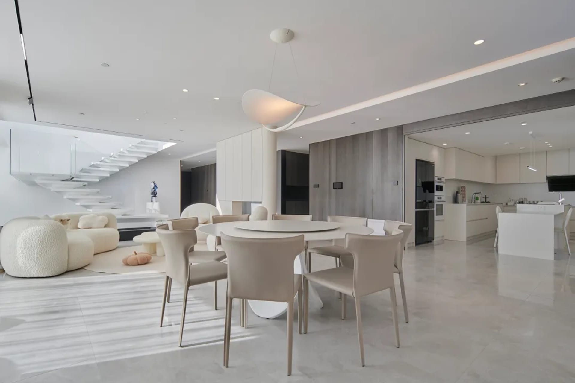 A modern living, dining, and kitchen area with white marble floors, a white round dining table surrounded by beige chairs, an