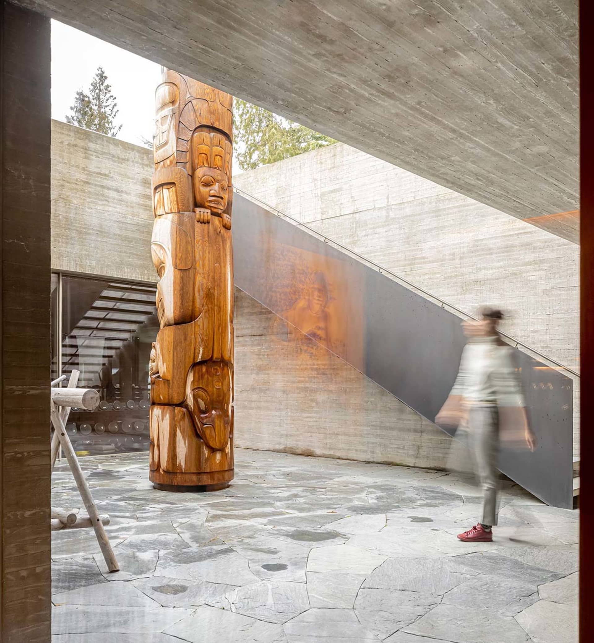 A modern indoor space featuring concrete decoration with a wooden totem pole, stone floor tiles, and a person walking on the 