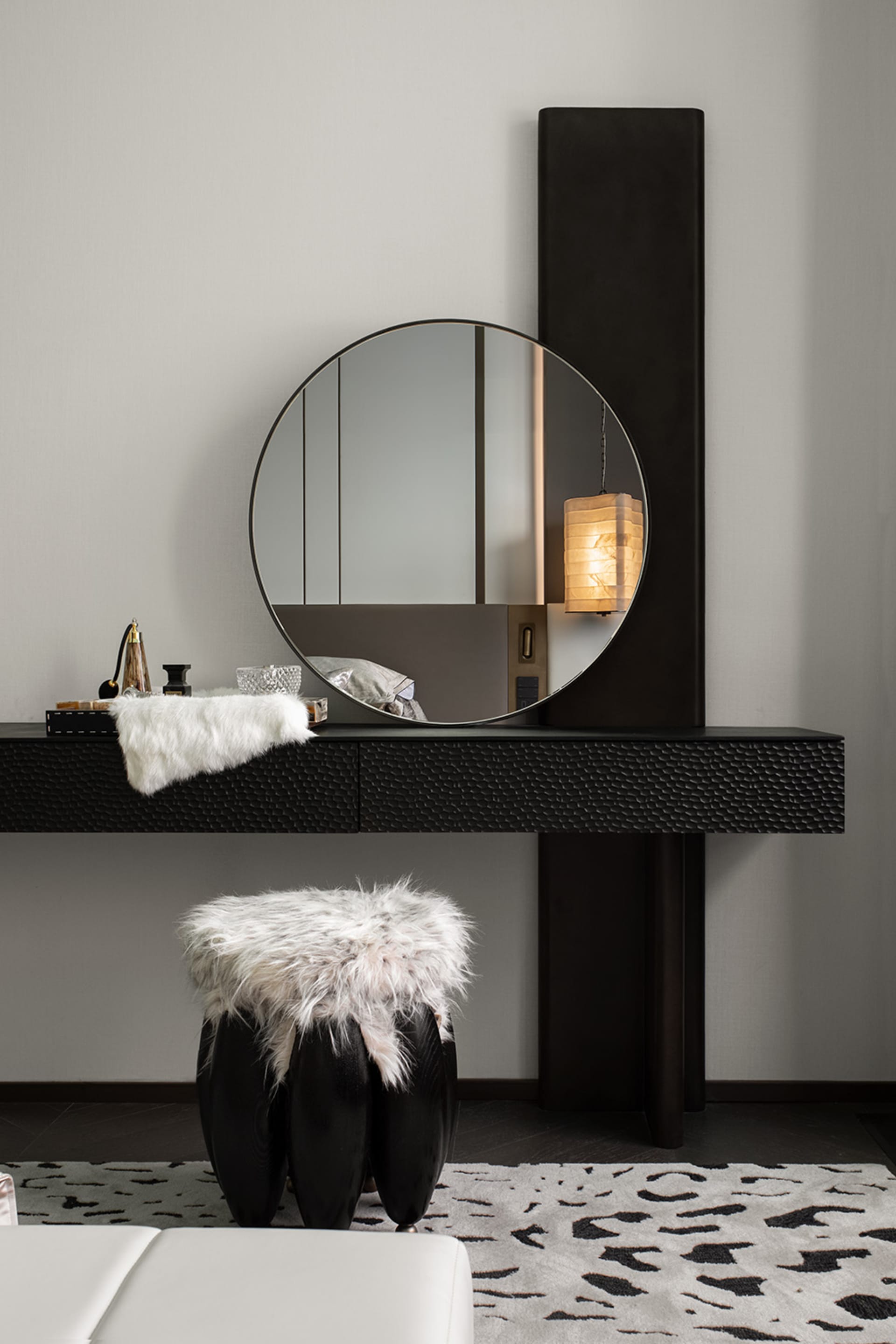 A modern interior design featuring a dressing table with a round mirror, black metal table, and a white faux fur stool on a p