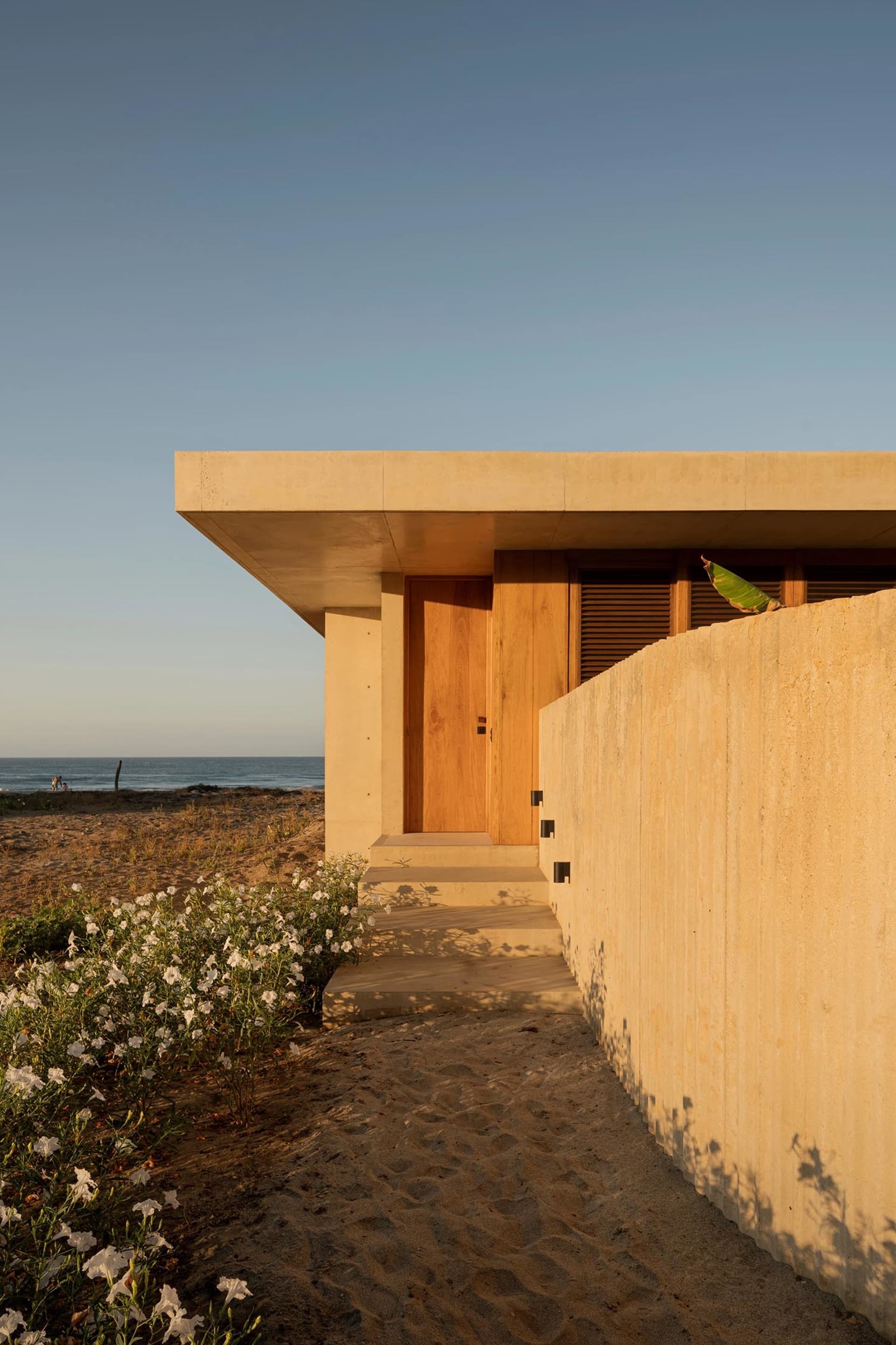 Modern minimalist residential exterior with concrete walls and a wooden door on a sandy beach.