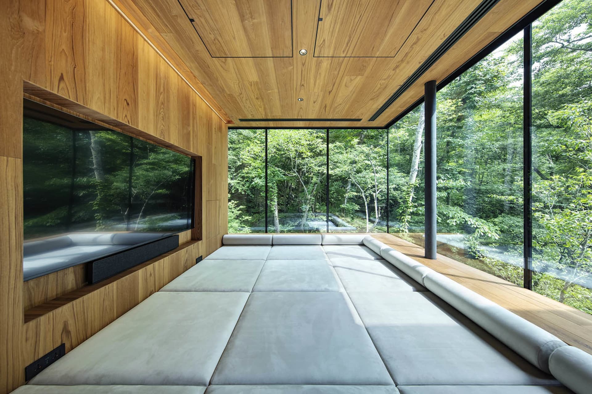 Modern living room with plush seating, wooden walls, and large glass windows overlooking a forest.