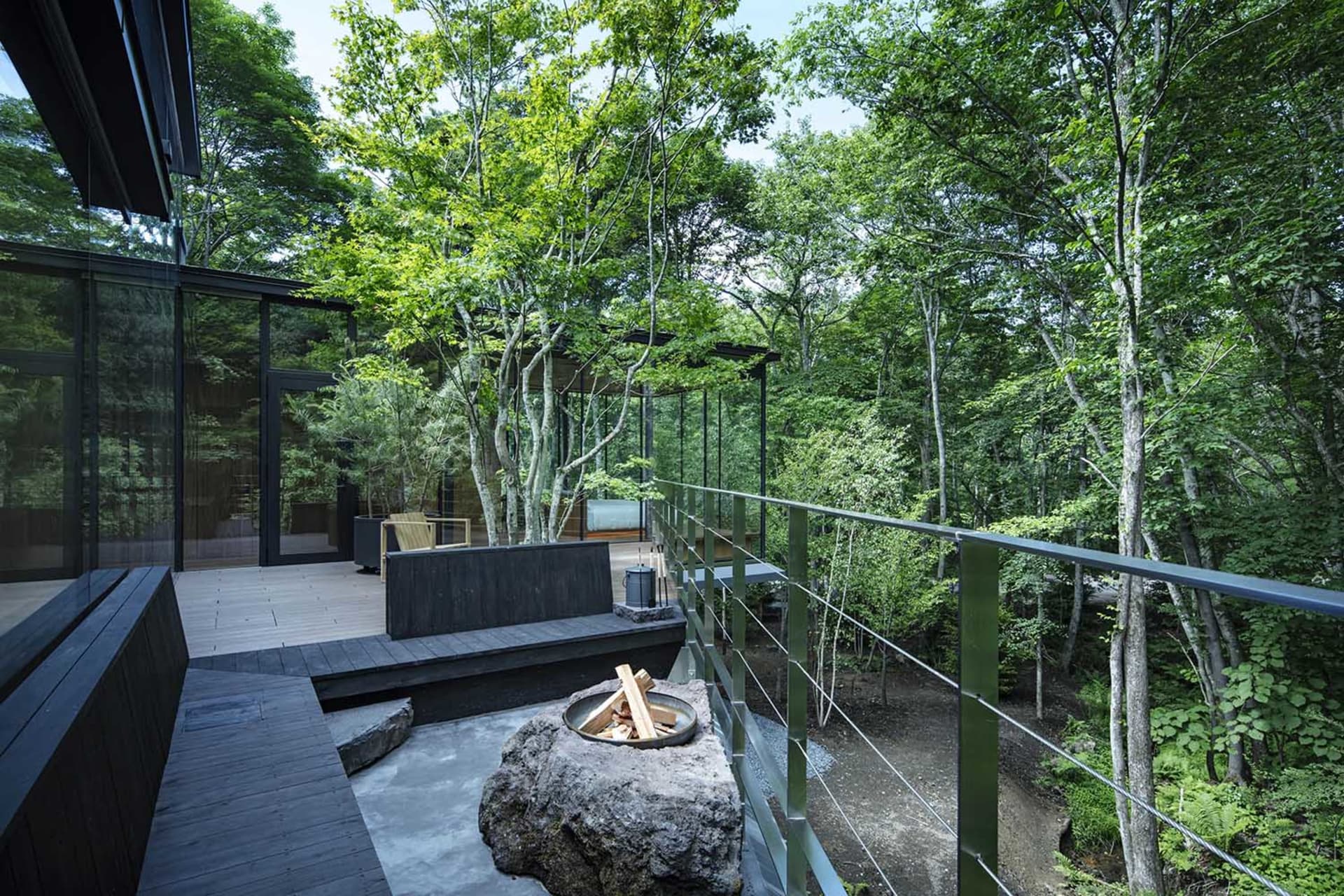 A modern outdoor space featuring a wooden deck, glass railings, and a concrete fire pit surrounded by lush greenery.