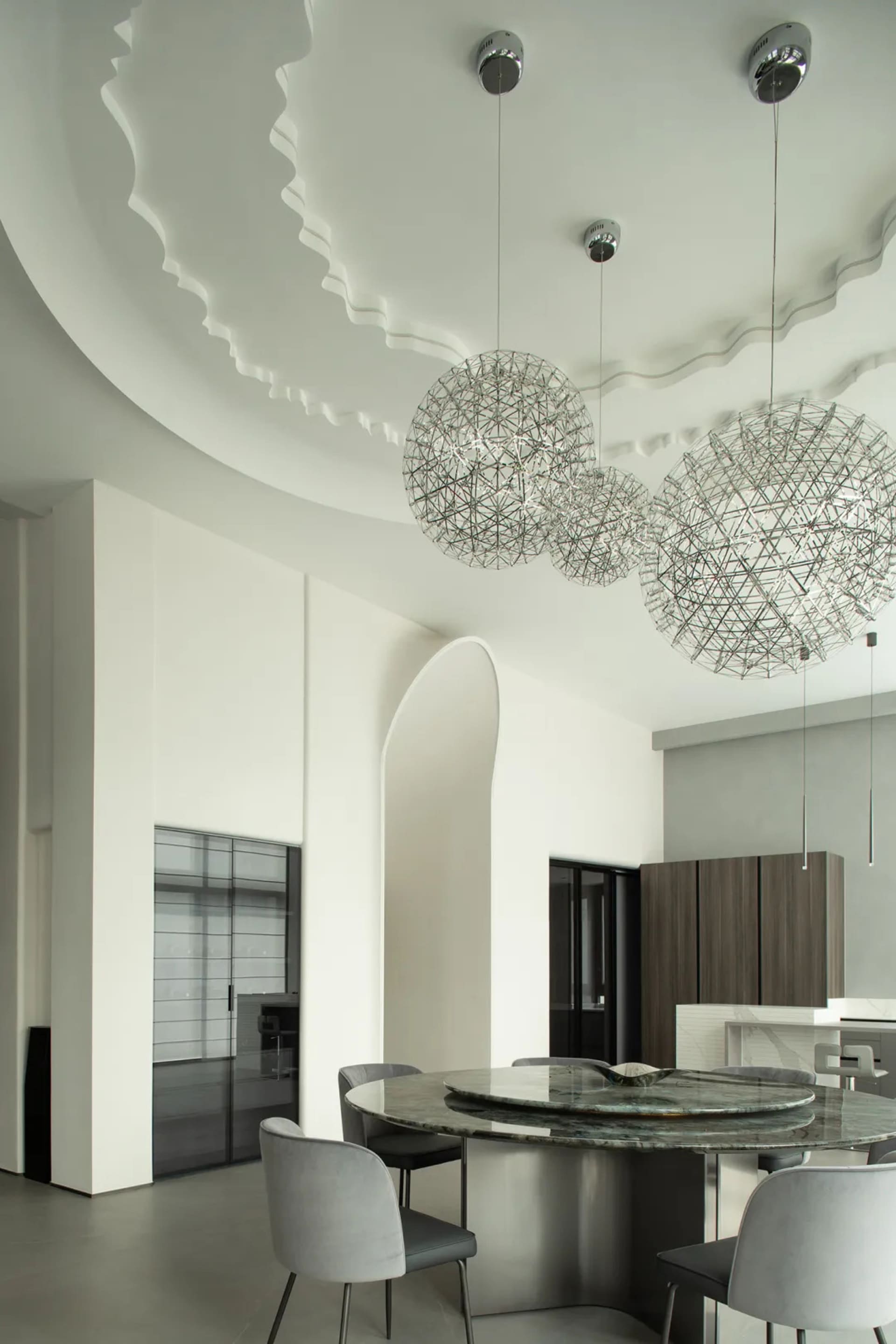 Modern dining room featuring a metal chandelier with geometric patterns, grey upholstered chairs, and a round granite table.