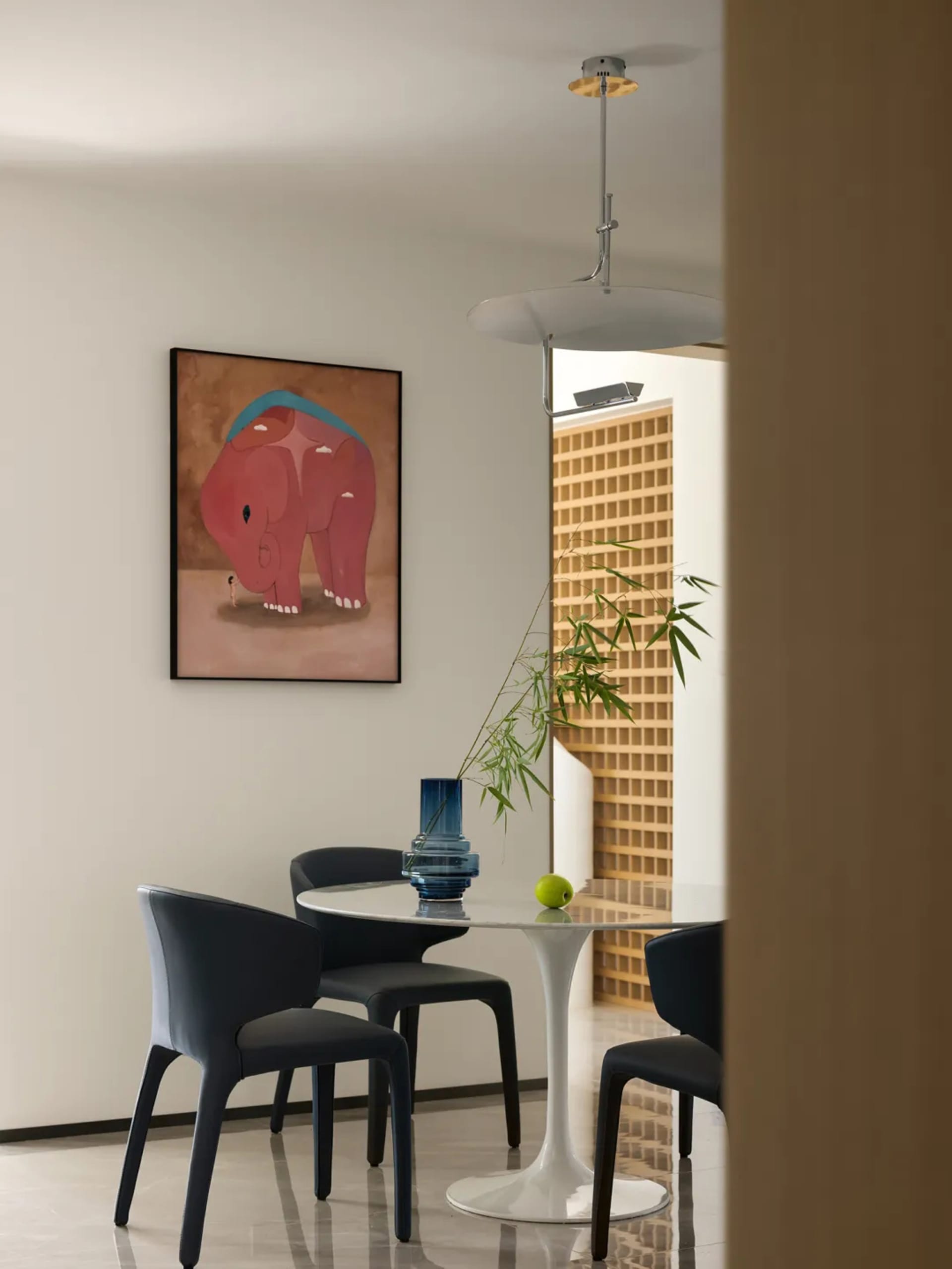 A minimalist dining room with a white round table, blue chairs, and a blue vase with greenery and an apple. A painting of a r