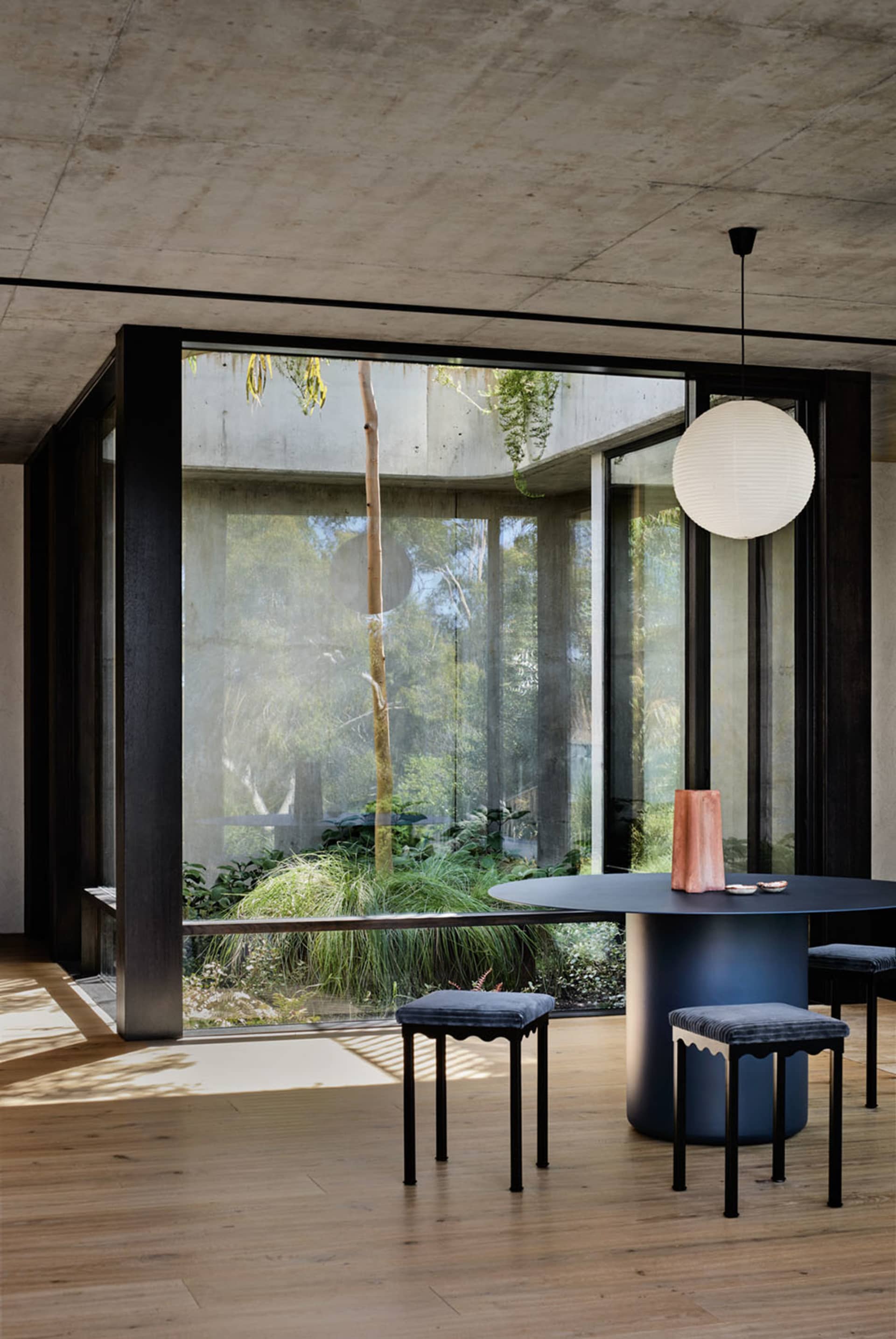 A modern, minimalist dining room with a large round table, stools, and a concrete and glass structure visible through large w