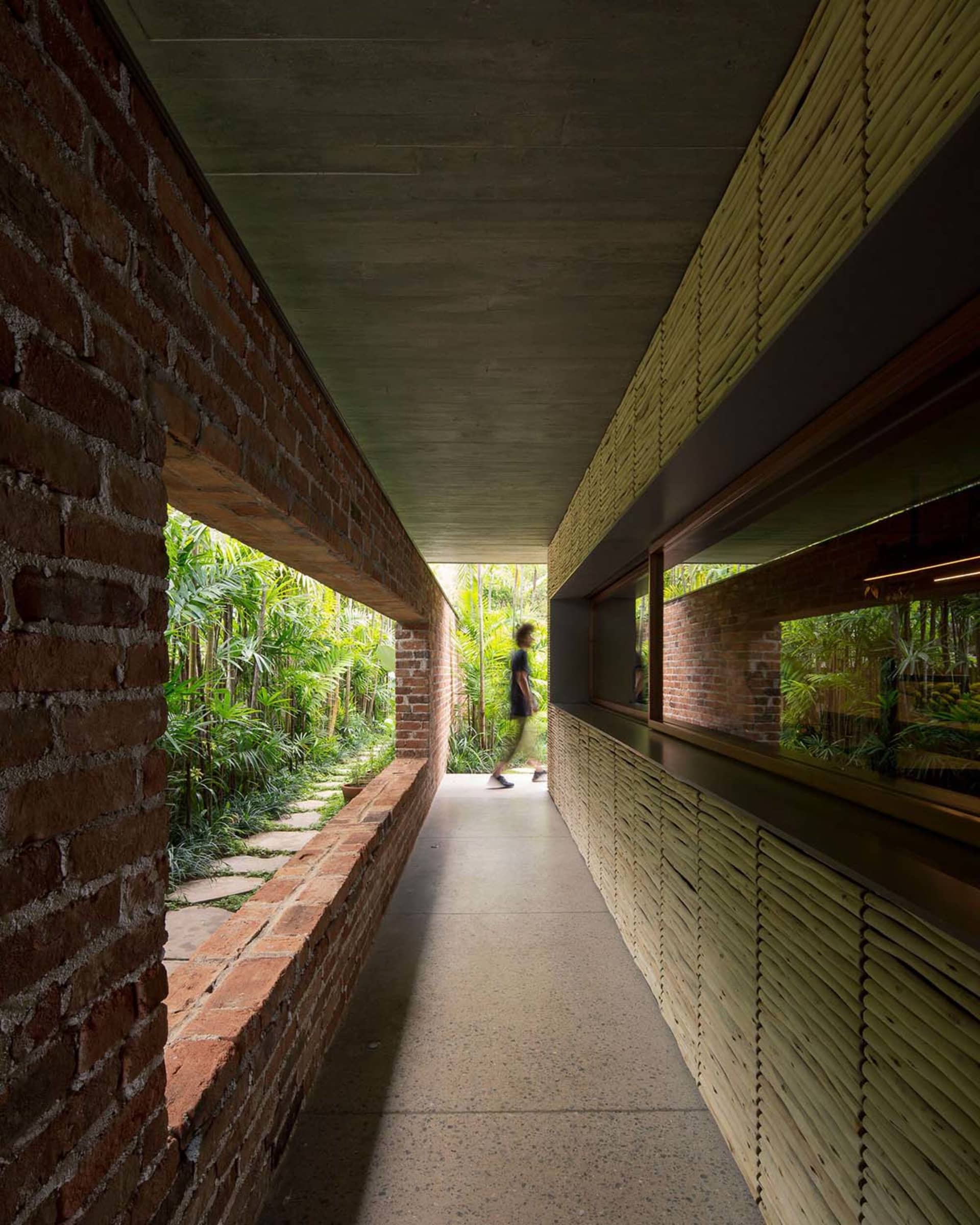 A modern industrial hallway features exposed brick and concrete walls, with a person walking along the path lined with tropic