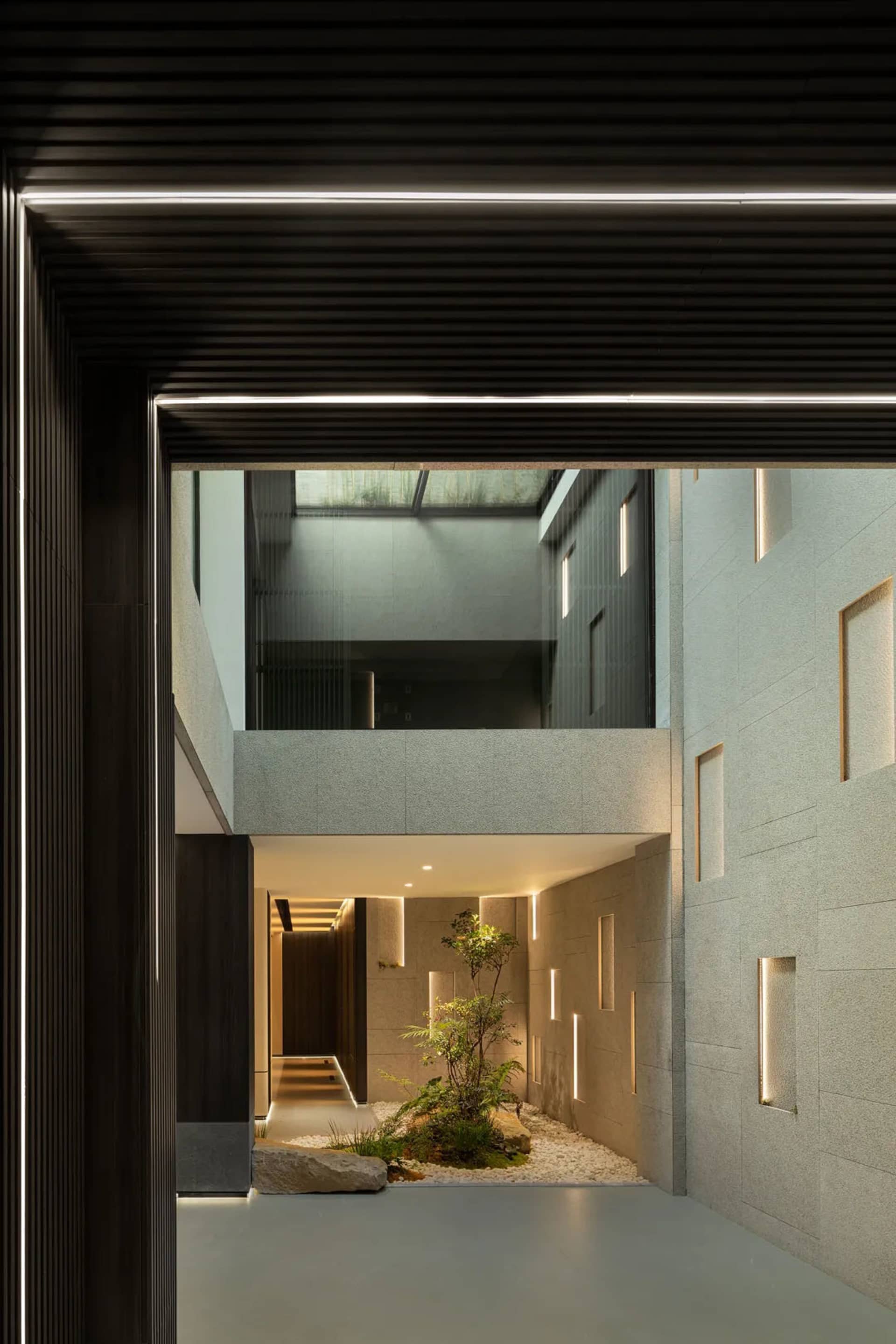 A modern residential entryway featuring concrete and glass materials, with a small indoor garden and illuminated by subtle li