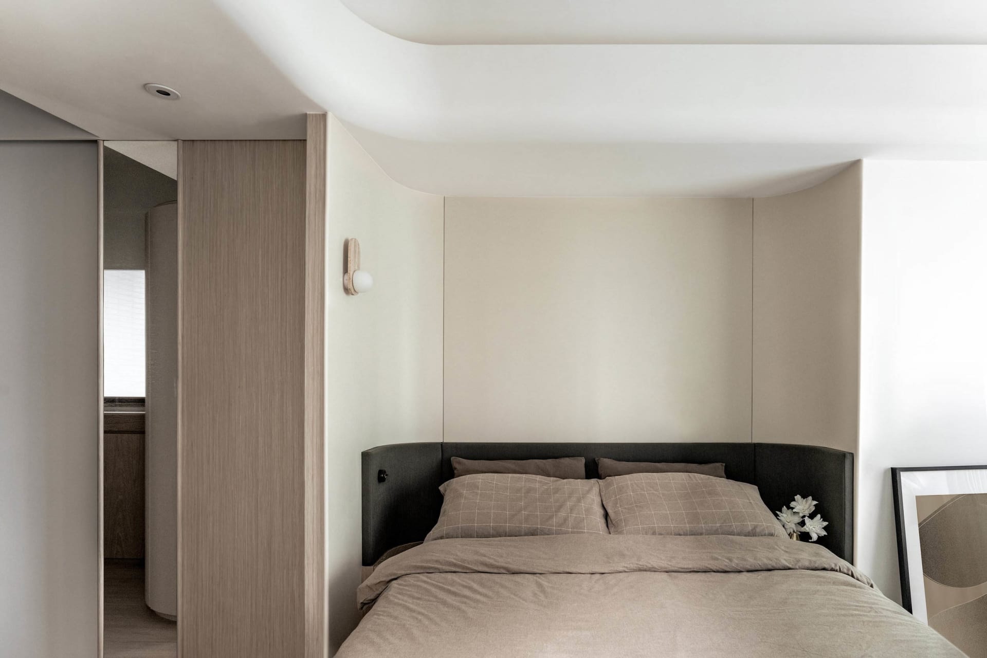 A minimalist bedroom featuring a bed with a black upholstered headboard and beige bedding, alongside a wooden headboard panel