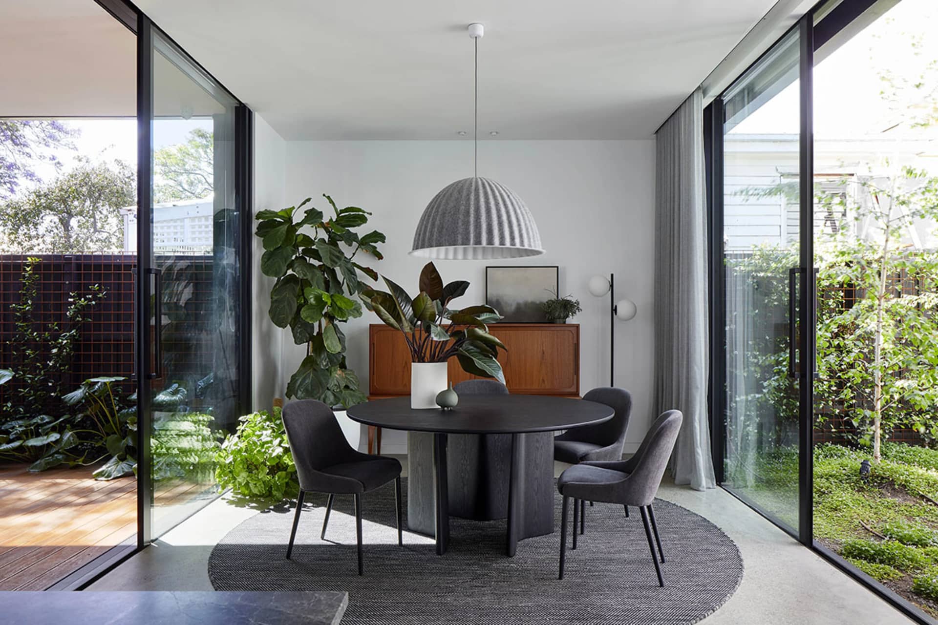 A modern dining room with a round black dining table, black upholstered chairs, a large green plant, and a round pendant ligh