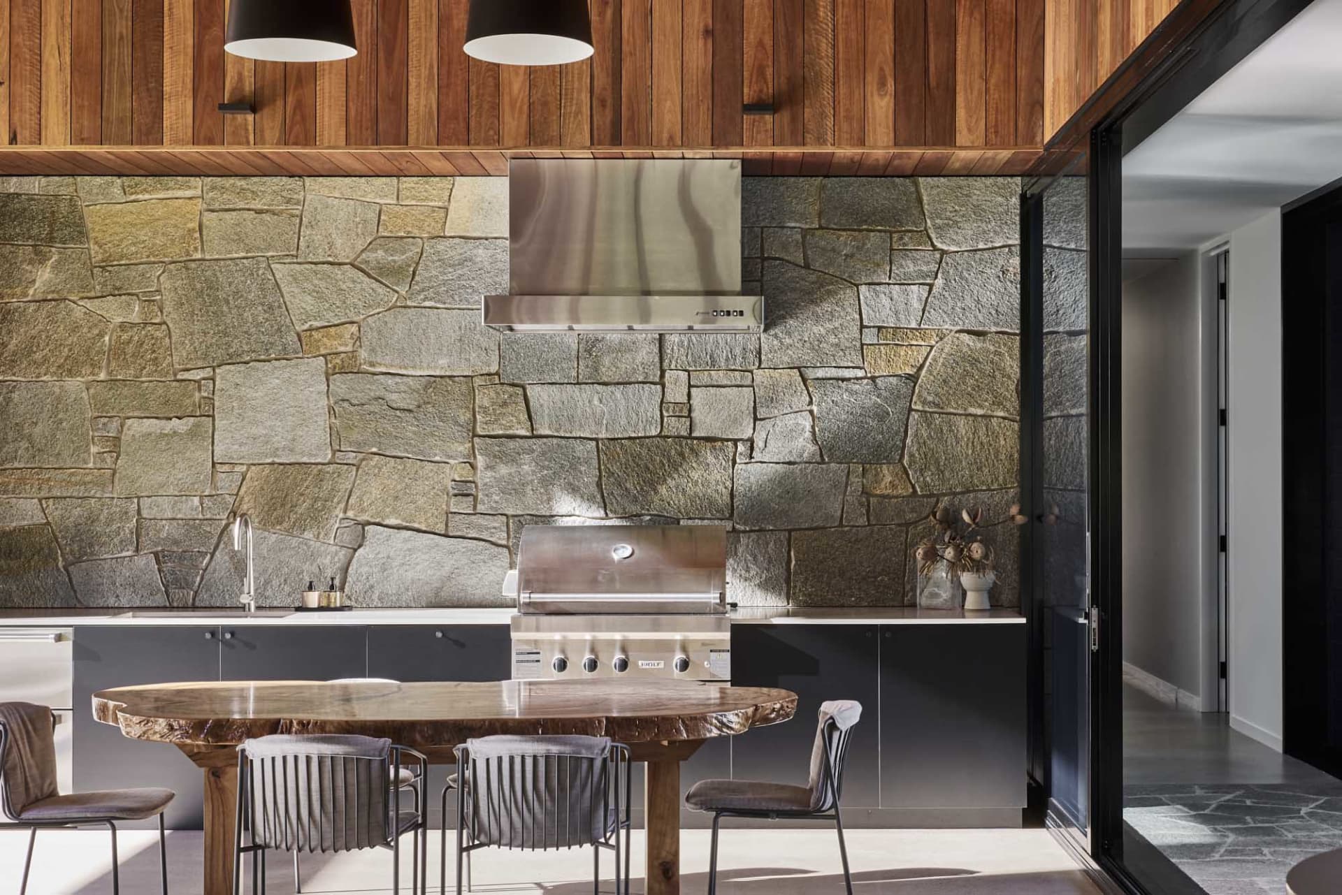 A modern kitchen with stone and concrete walls, a teak wood table, and a stainless steel grill and hood.