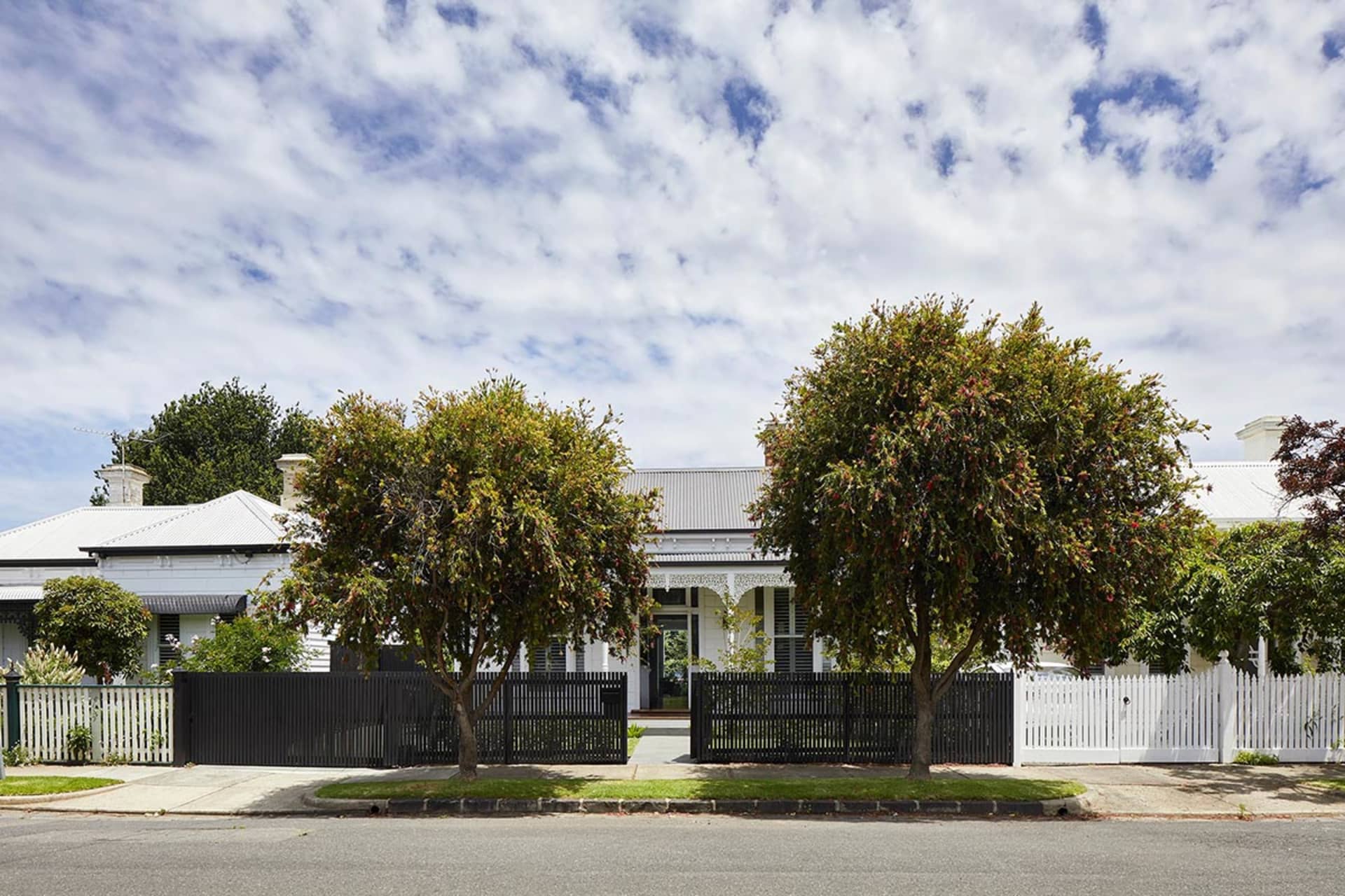 A white Victorian-style house with a brick fence and trees in front of it, set against a blue sky with white clouds.
