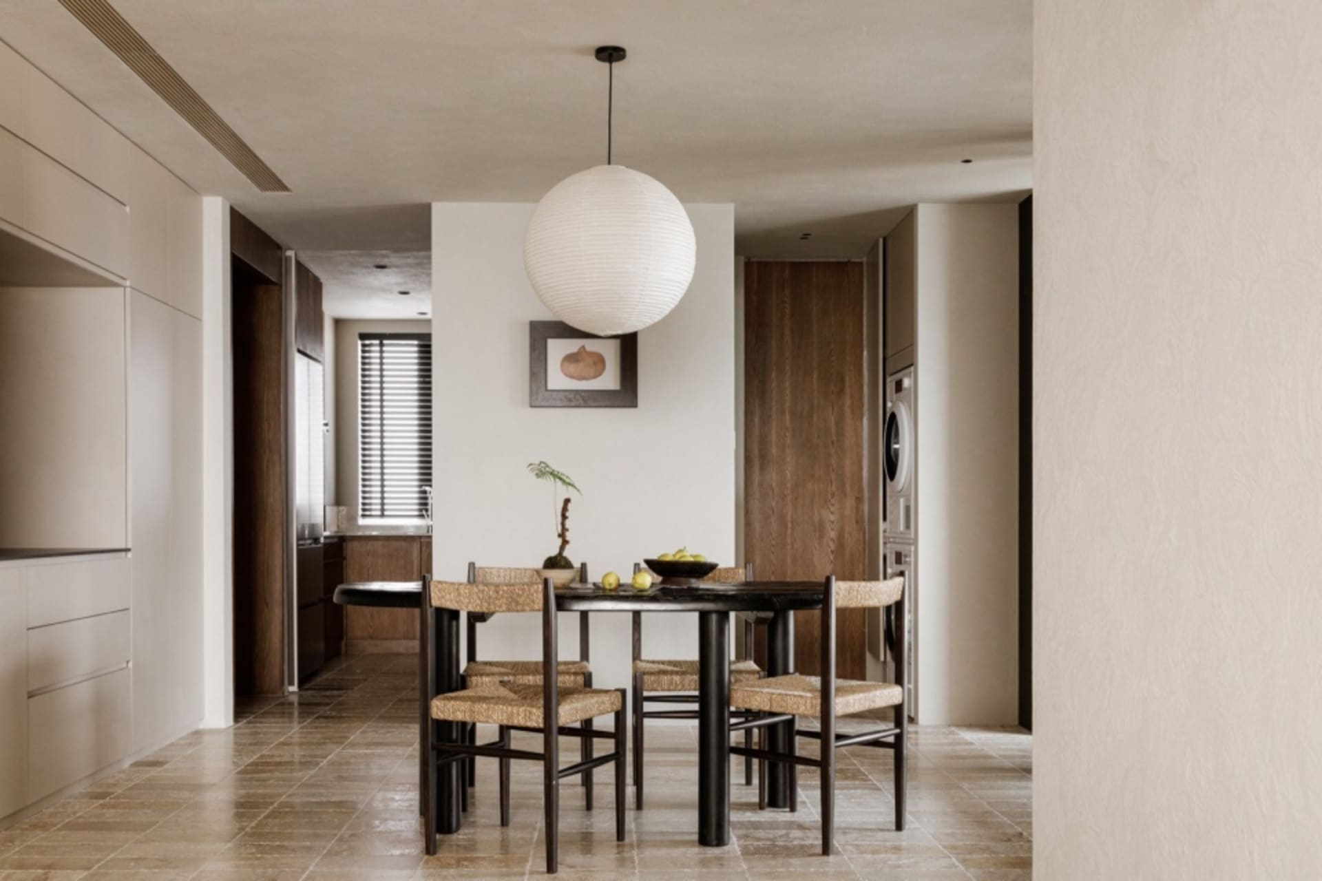 A modern, minimalist kitchen and dining room featuring a black dining table surrounded by woven dining chairs and marble coun