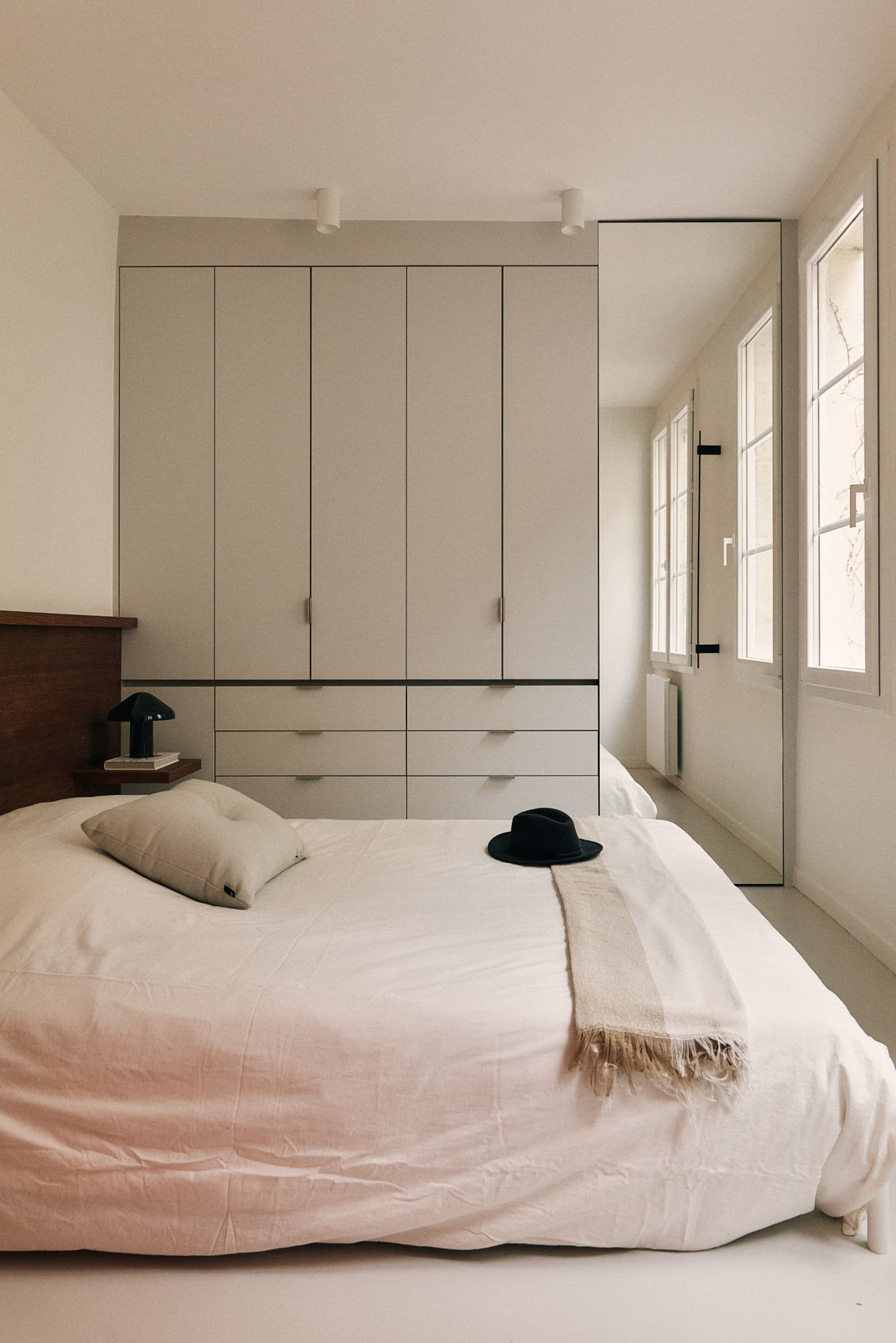 A modern minimalist bedroom with concrete floors, white bedding, and a large window allowing natural light to flood the space