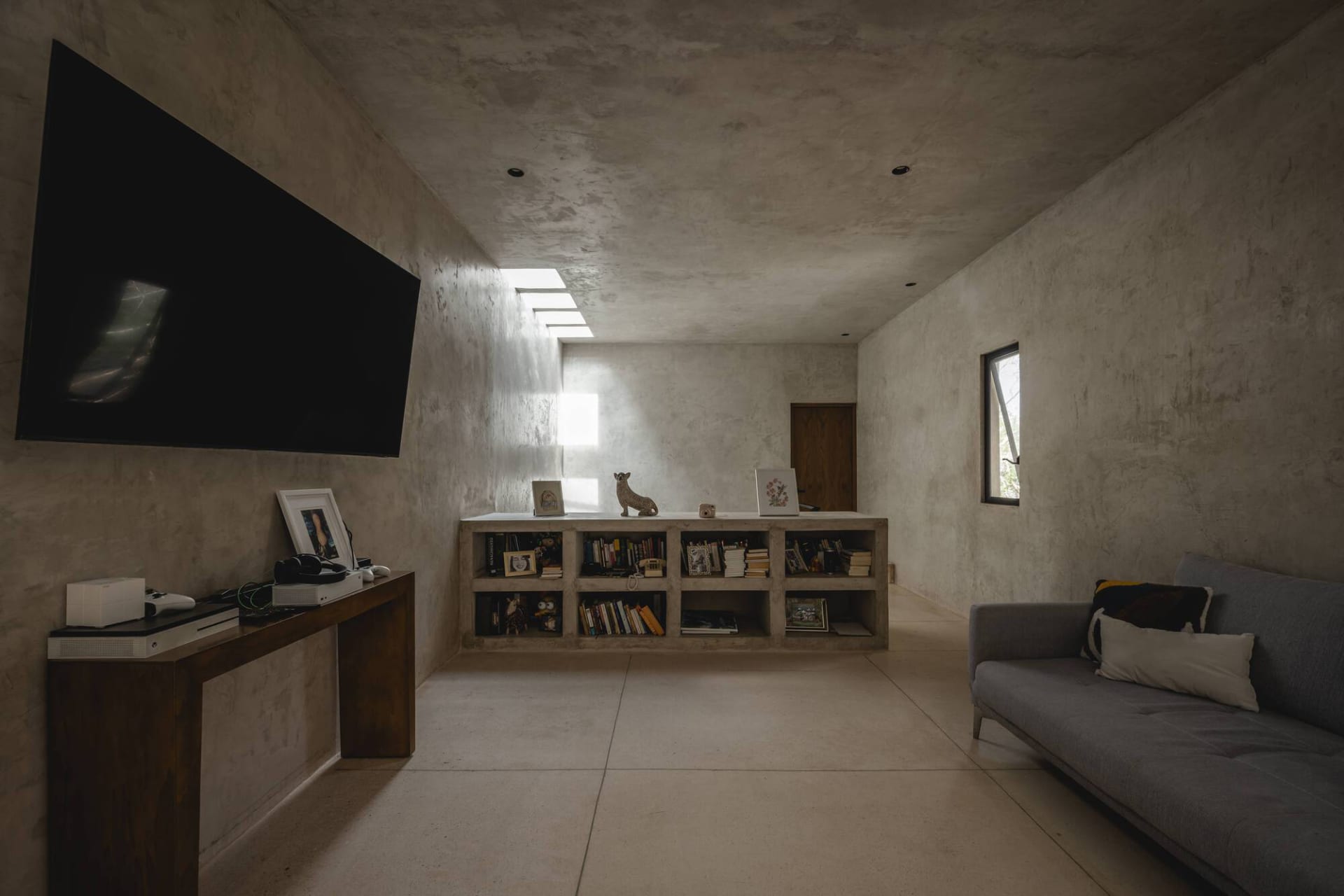 Modern minimalist living room with concrete walls, a low-slung bookshelf, a grey sofa, and a large screen TV mounted on the w
