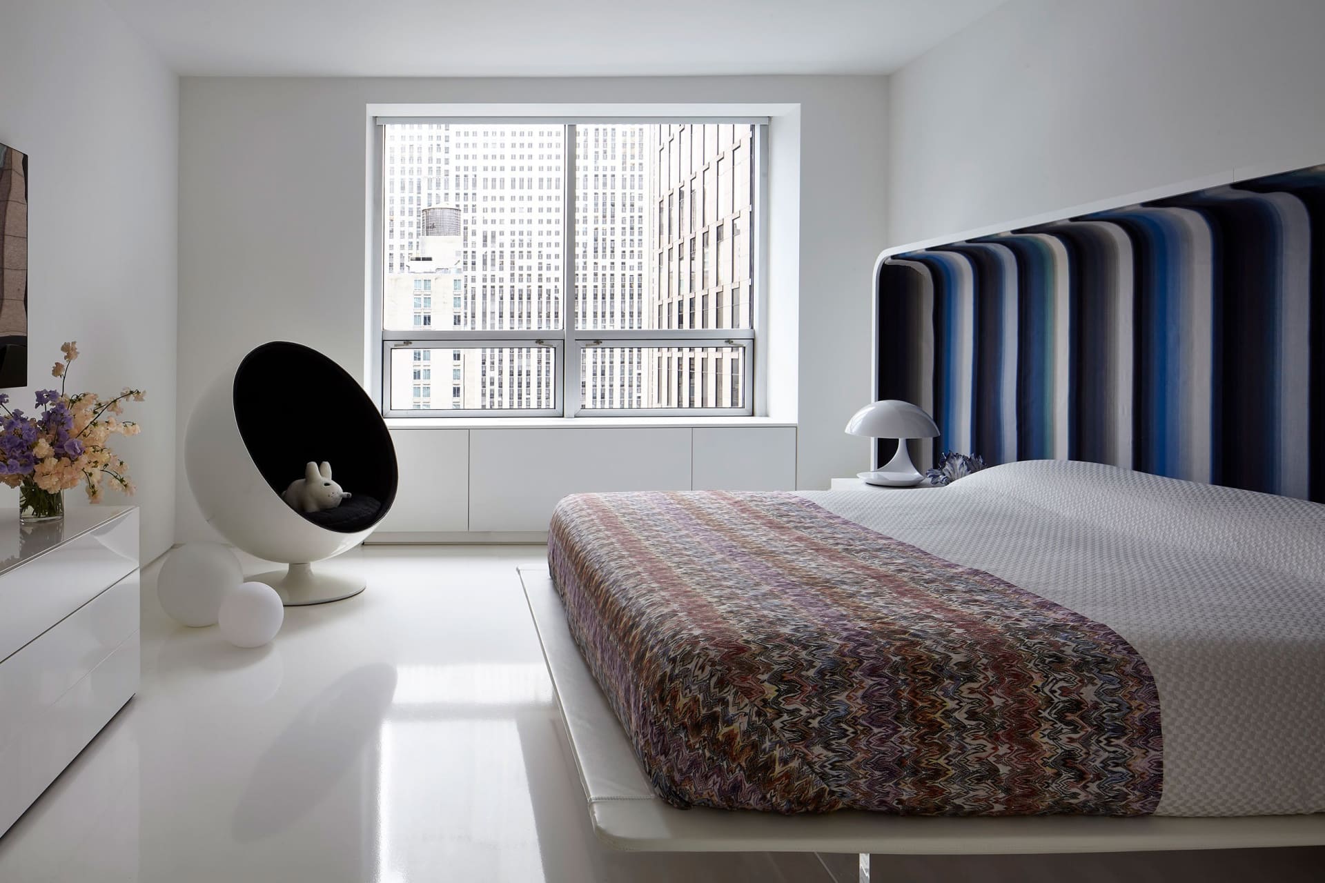 A minimalist bedroom featuring a modern bed with a colorful striped headboard and a sleek, white floor and furniture.