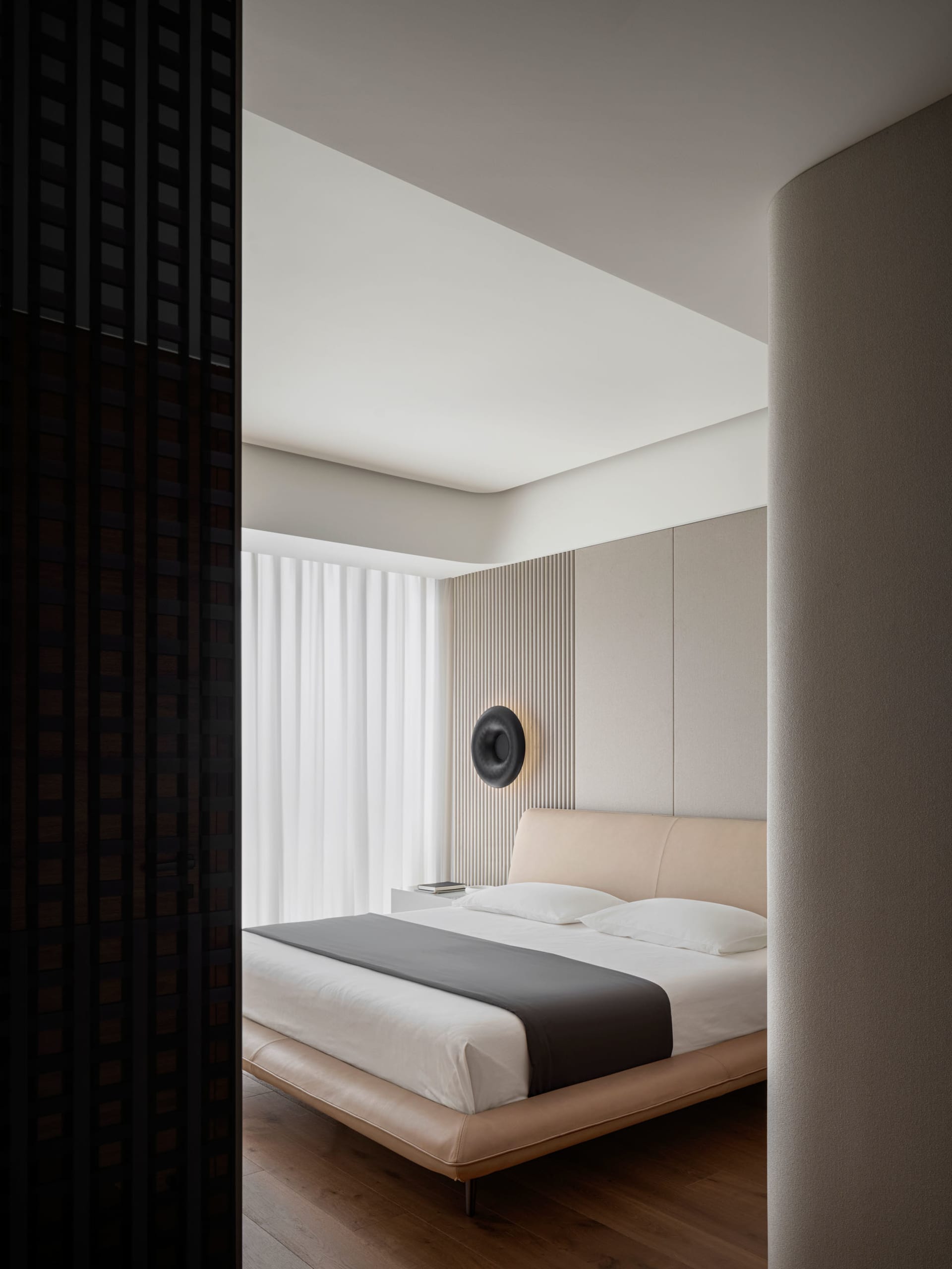 A minimalist bedroom with a beige upholstered bed, wooden flooring, and a round sconce light on the wall.