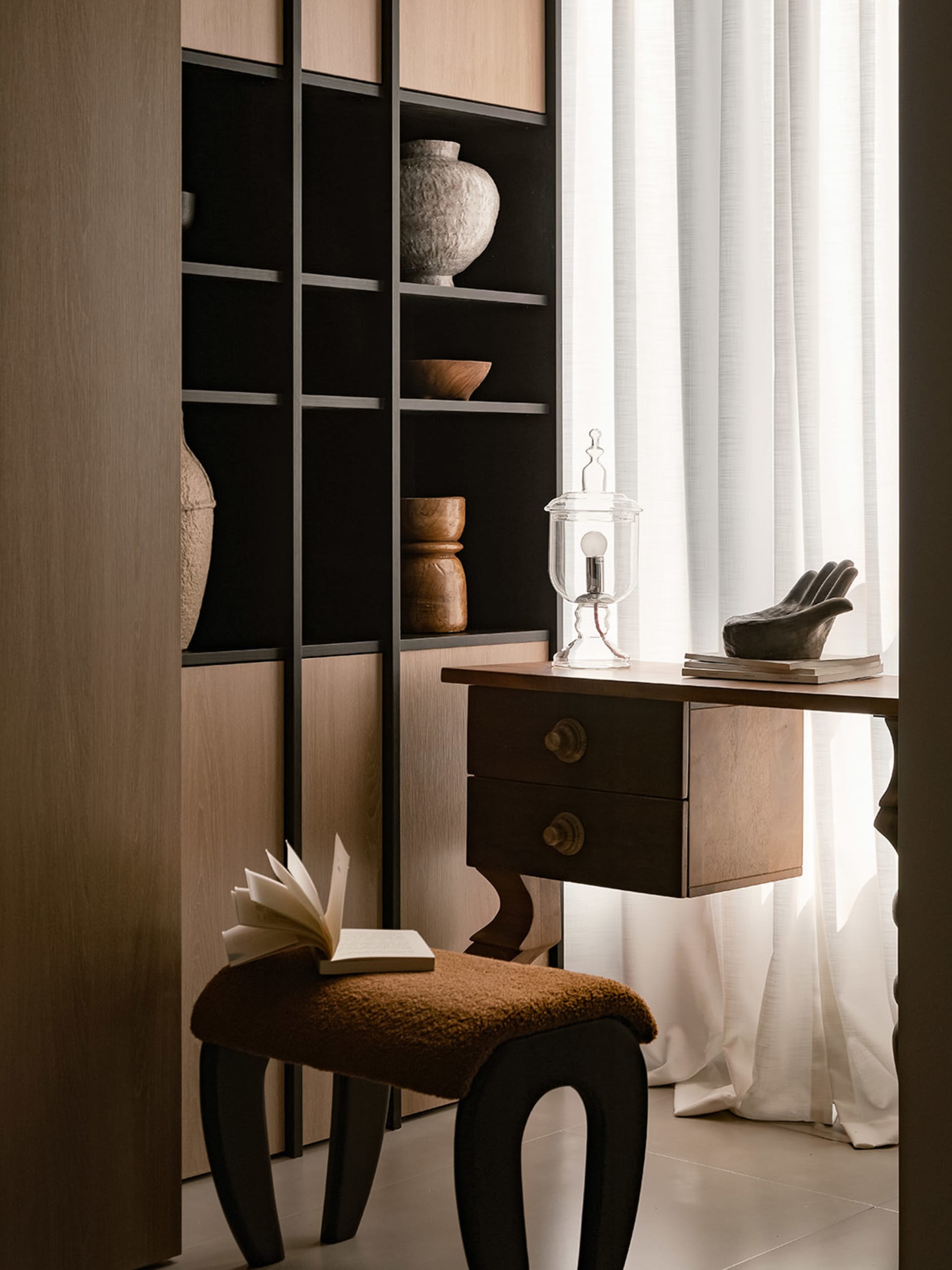 A minimalist home office featuring a wooden desk with a glass lamp and a book, a brown cushioned stool, and a bookshelf with