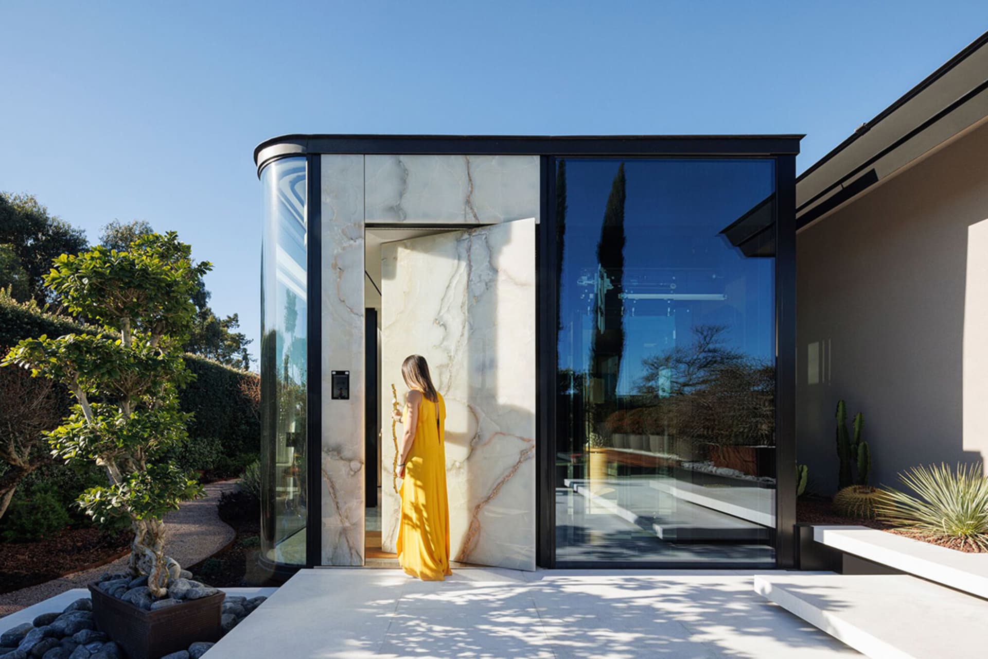 Woman in a yellow dress standing by a modern minimalist marble entrance with large glass panels.