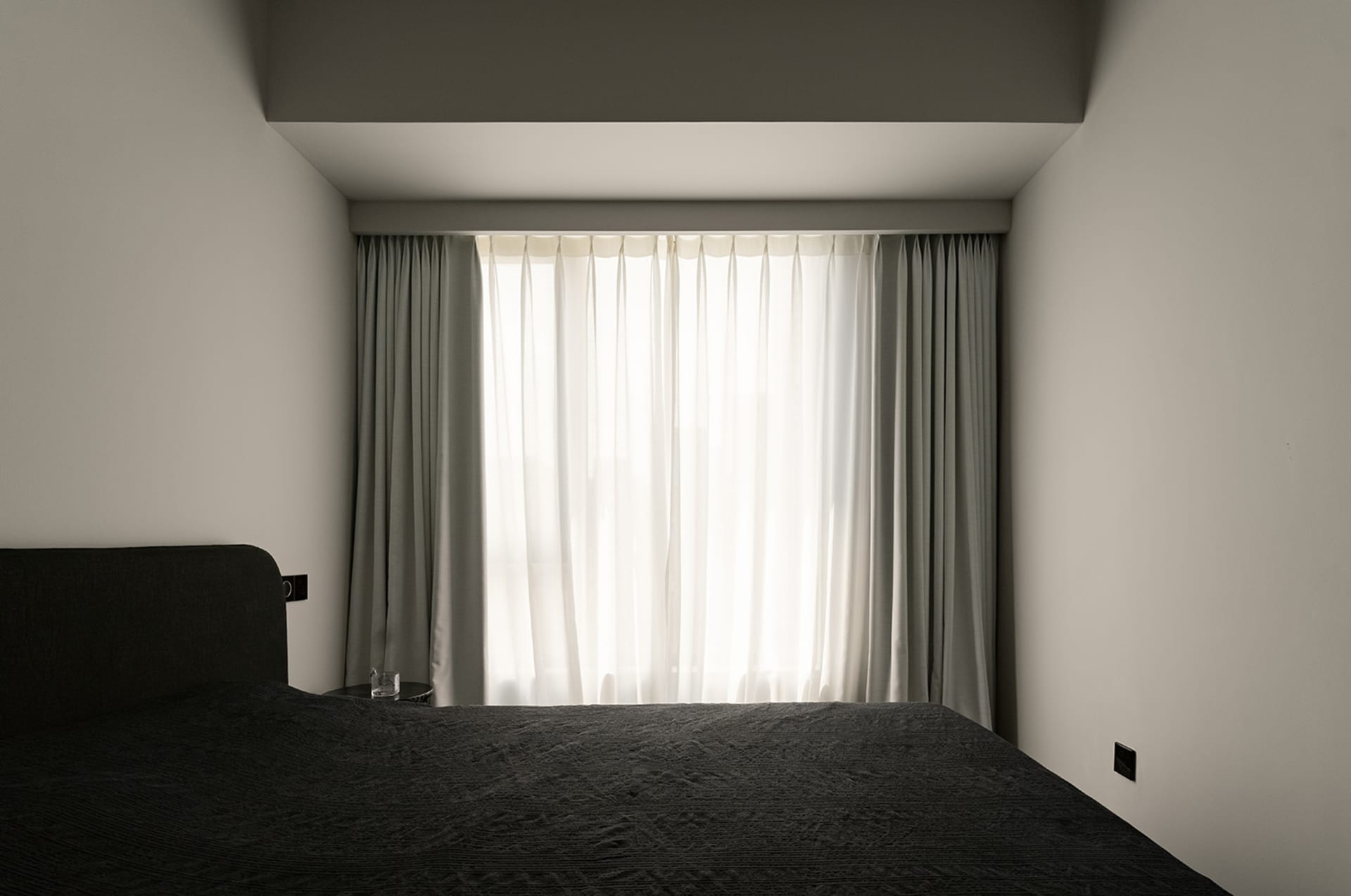 A minimalist bedroom with black furniture and grey walls, featuring white curtains and a window.