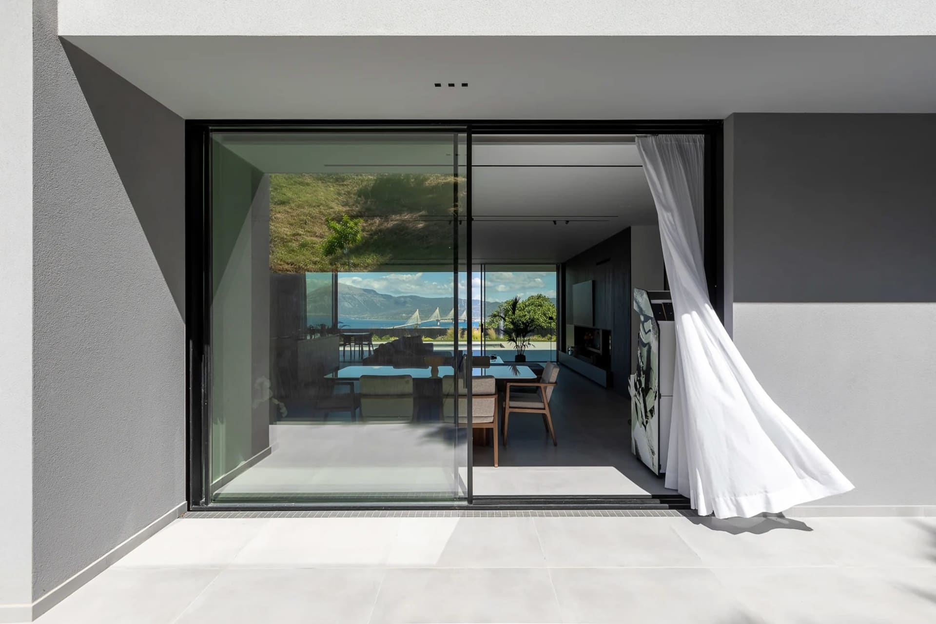 Modern minimalist living room with a glass and concrete patio overlooking a scenic view of mountains and water.