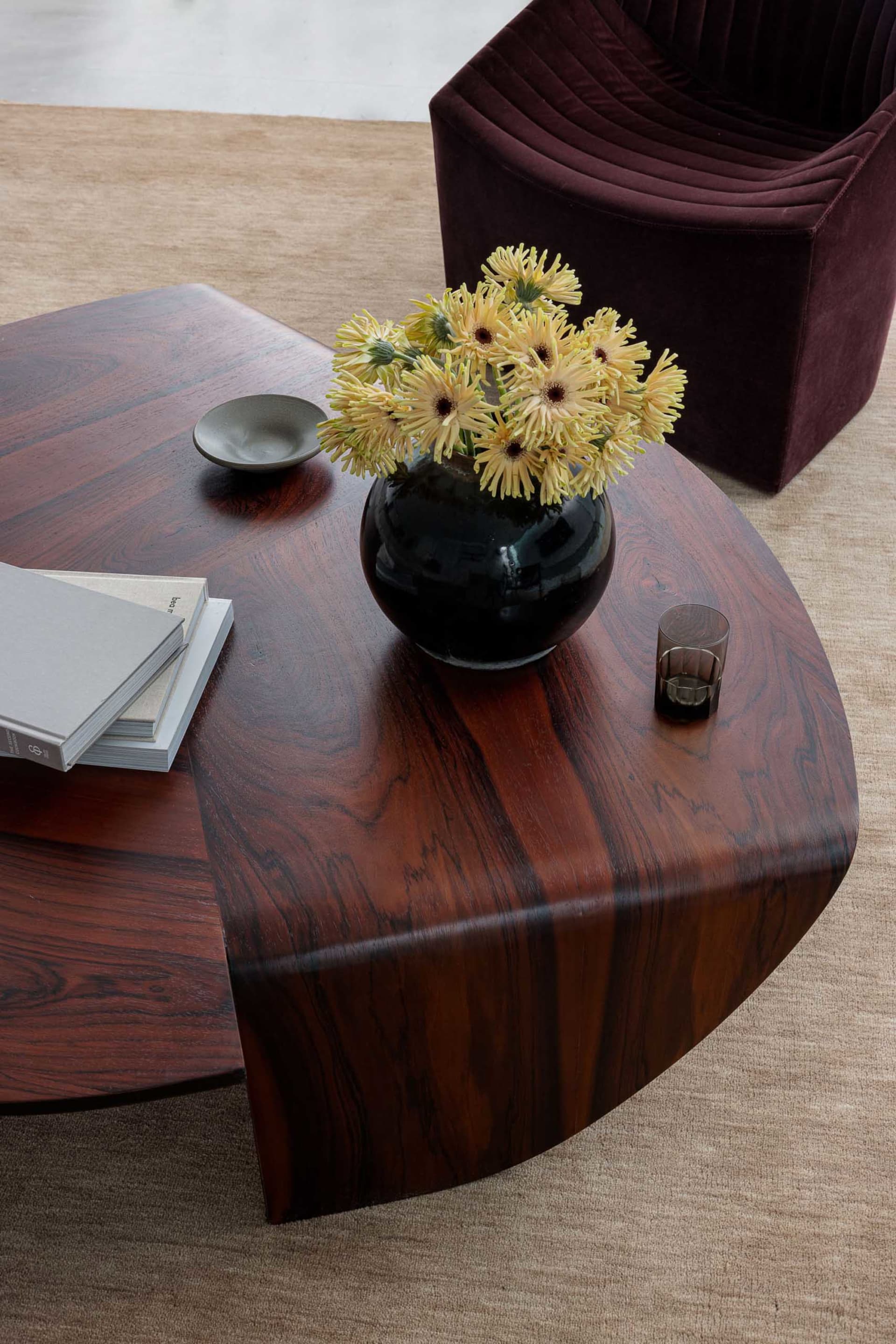 A modern coffee table setting featuring a dark brown wood veneer table with a black vase of yellow flowers, a stack of books,