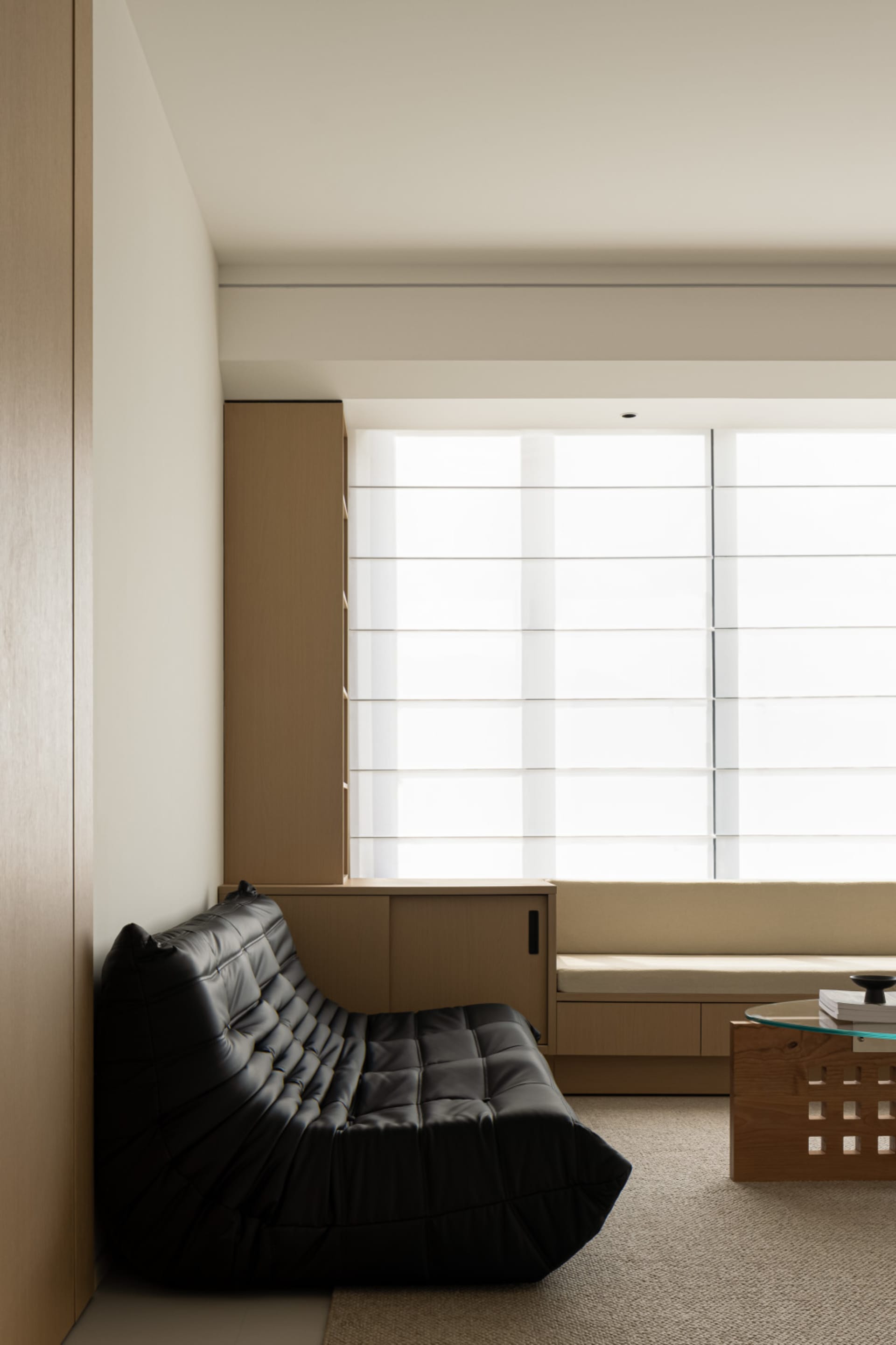 A modern living room with minimalist design, featuring a black leather sofa, wooden furniture, and large windows.