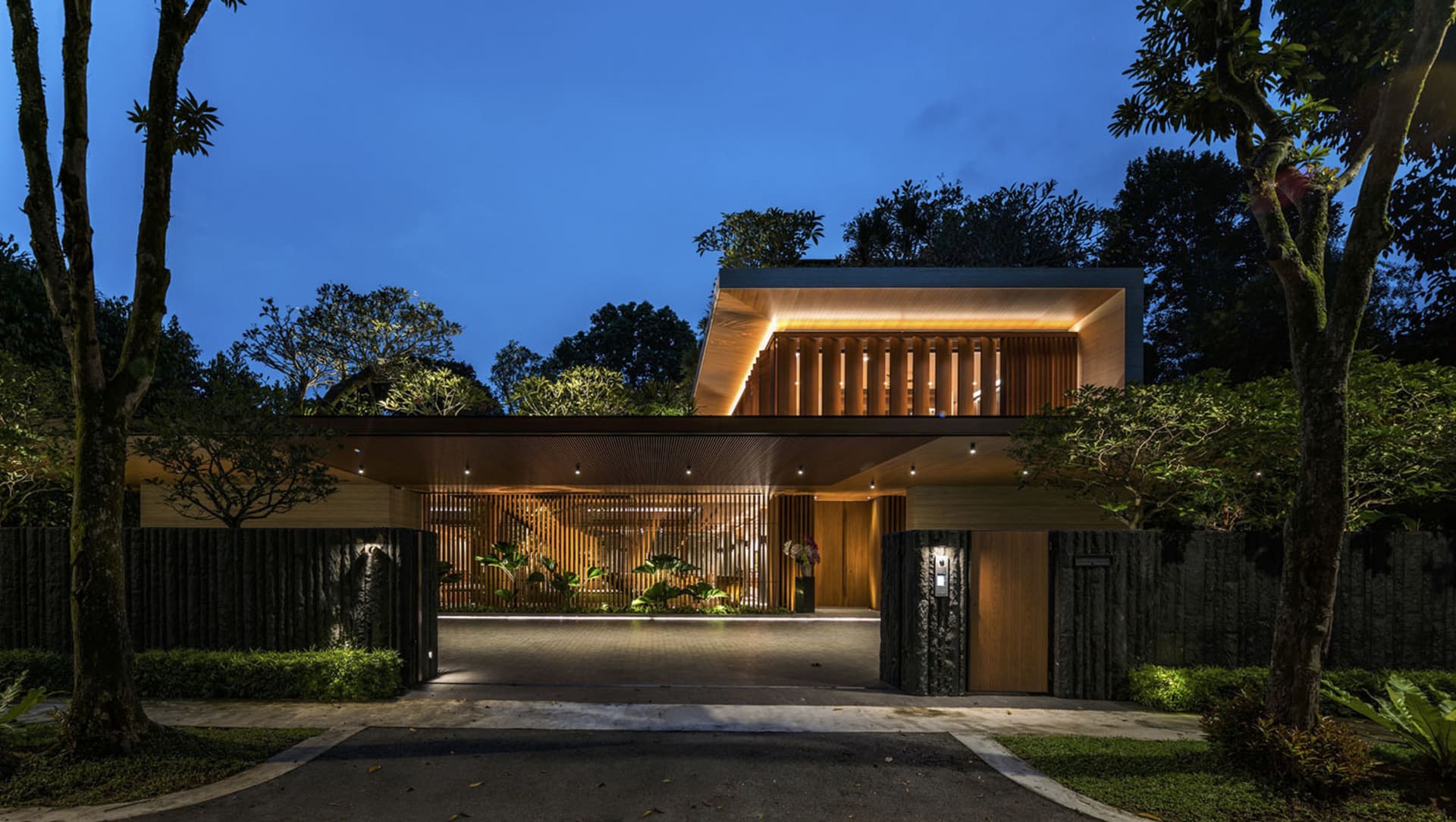Modern entrance with wooden panels of a residential house at dusk, featuring warm lighting and lush greenery.