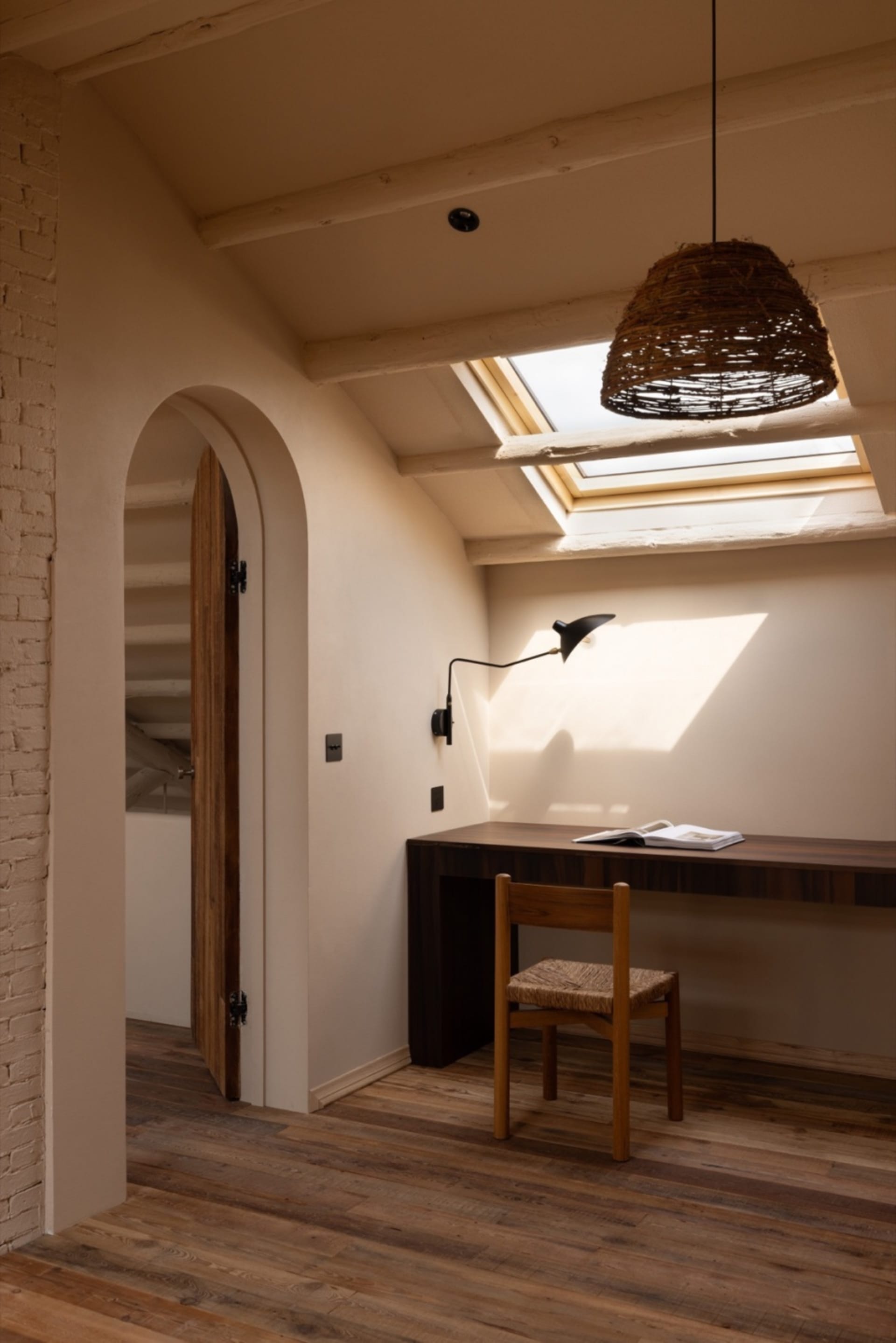 A minimalist home office with a timber floor, featuring a wooden desk, chair, and wicker pendant light under a skylight.