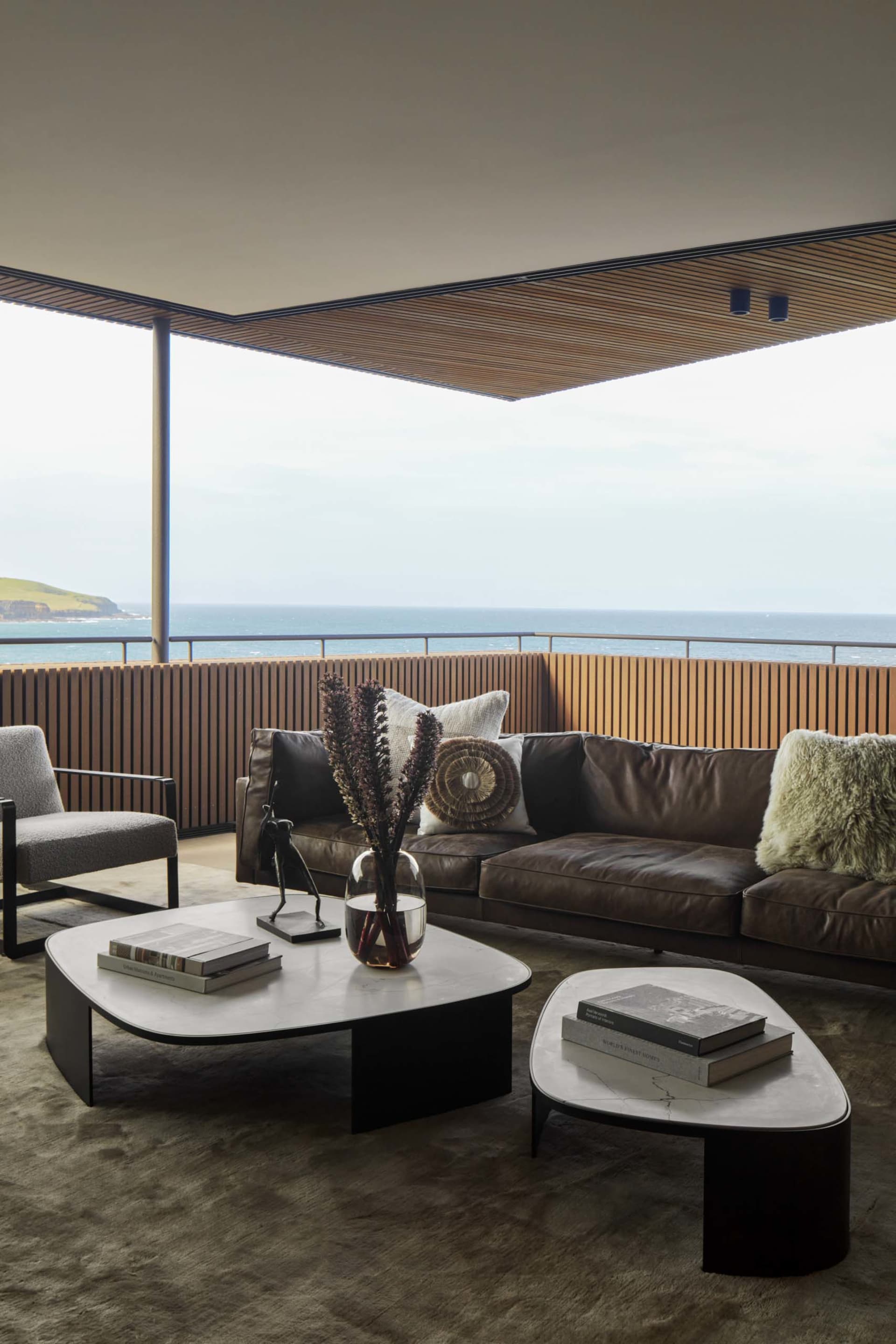 Modern industrial-style living room with a brown leather sofa, wooden coffee table, gray armchair, and large ocean view.