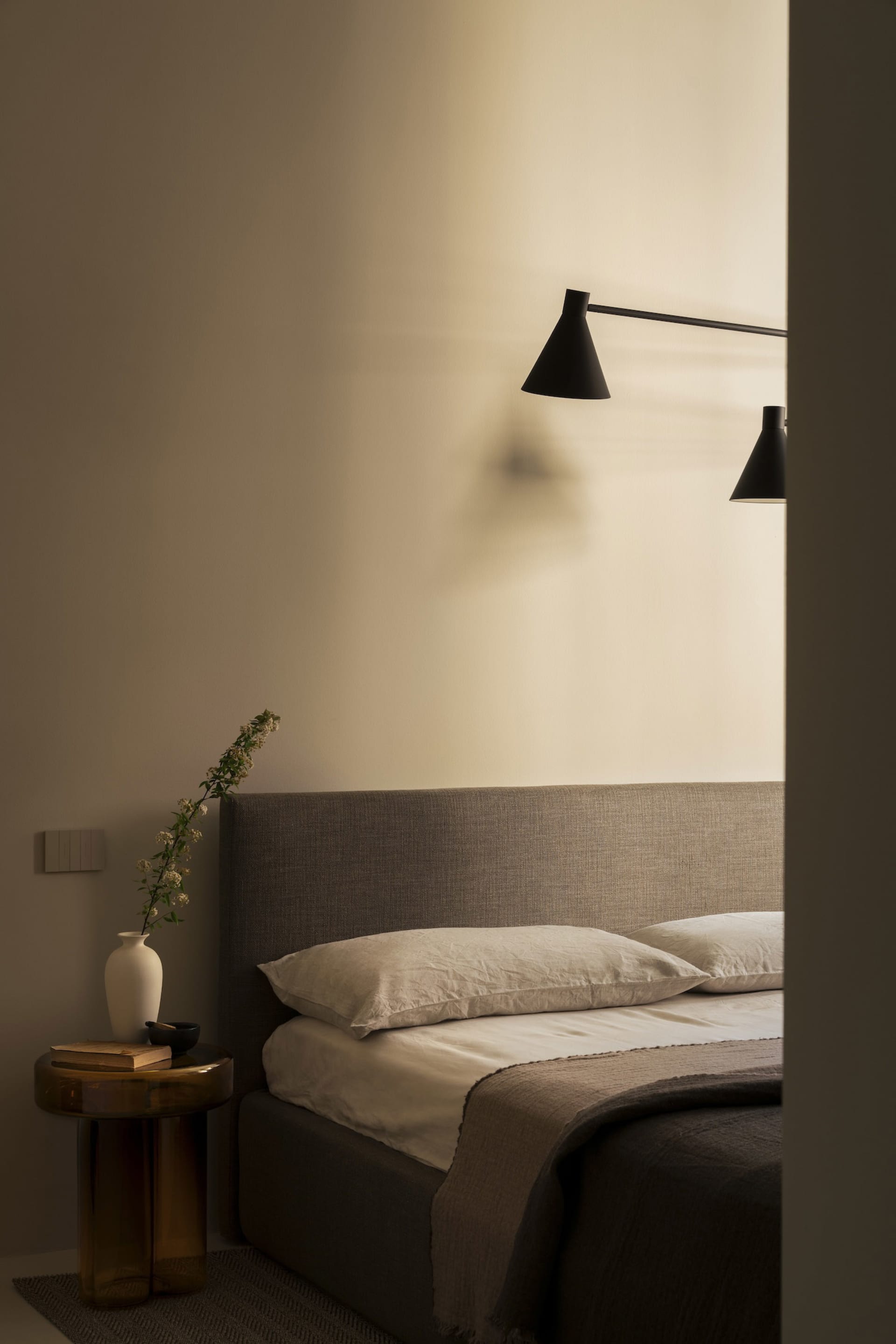A minimalist bedroom with a textured fabric headboard, two pillows, and a simple nightstand with a vase and flowers.