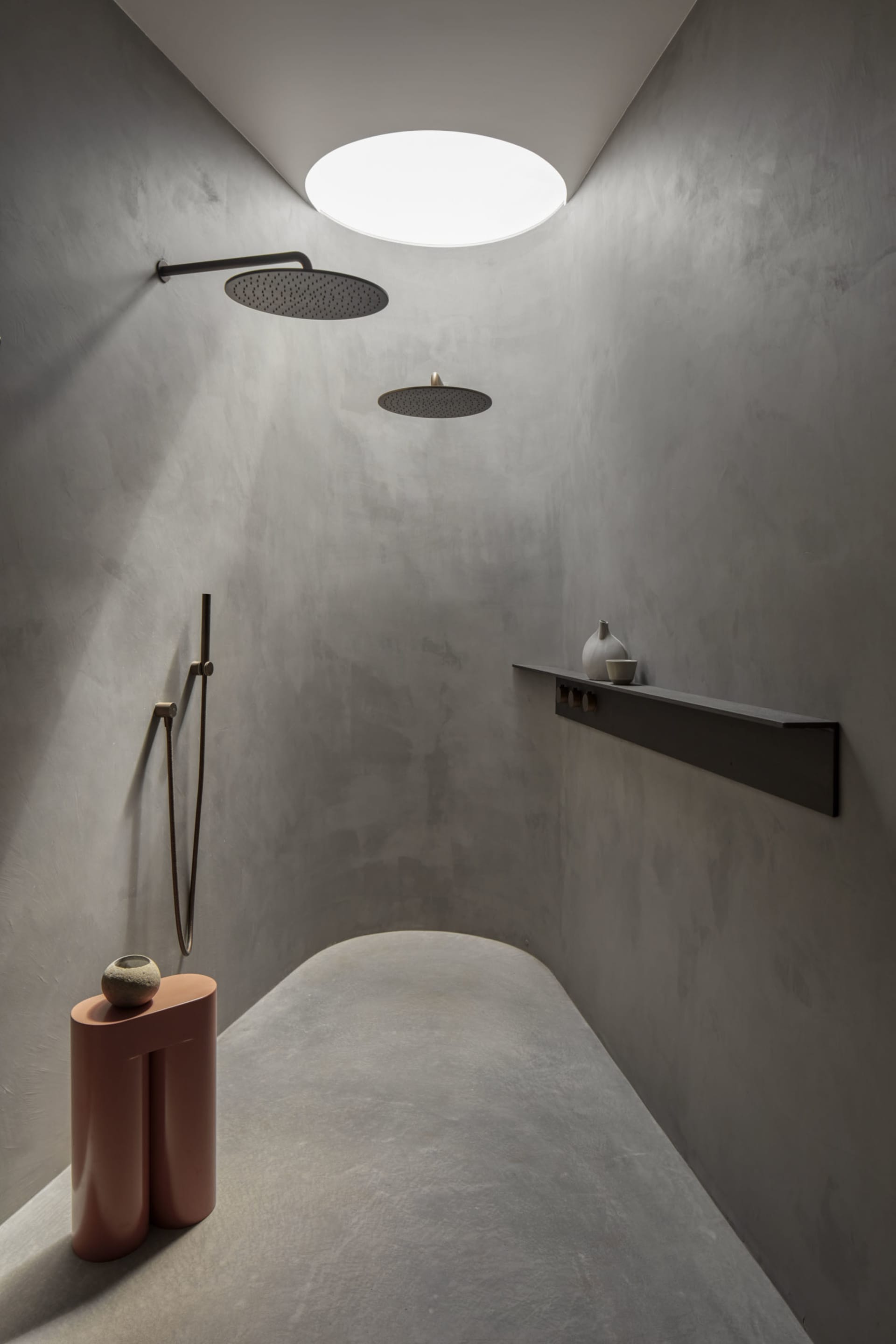 A minimalist bathroom with concrete walls, two overhead showers, and a small shelf with decorative items.