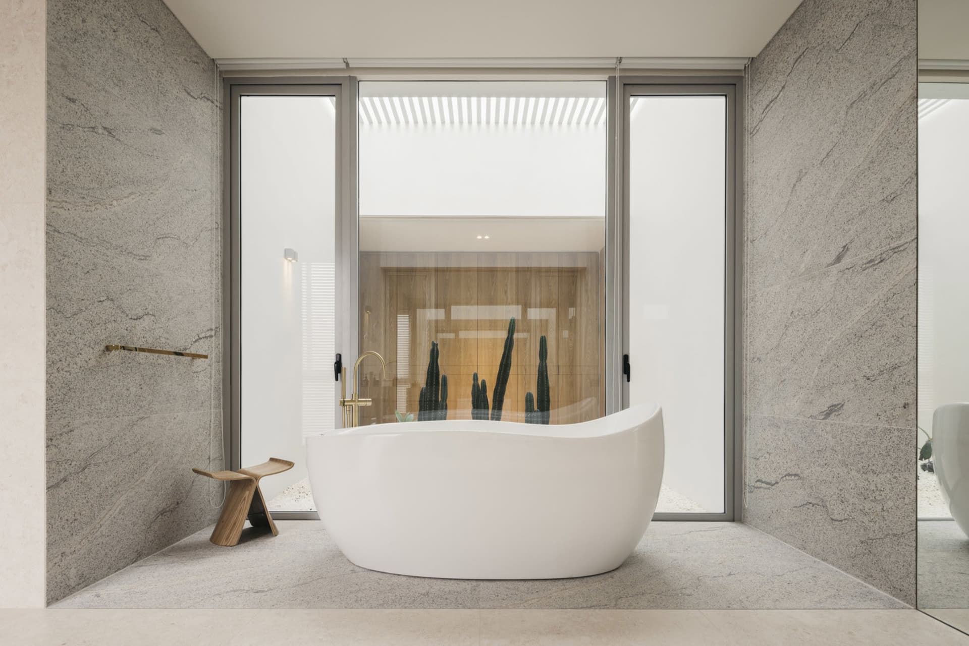 A minimalist bathroom with a white bathtub, wooden stool, and large windows showing a cactus plant.
