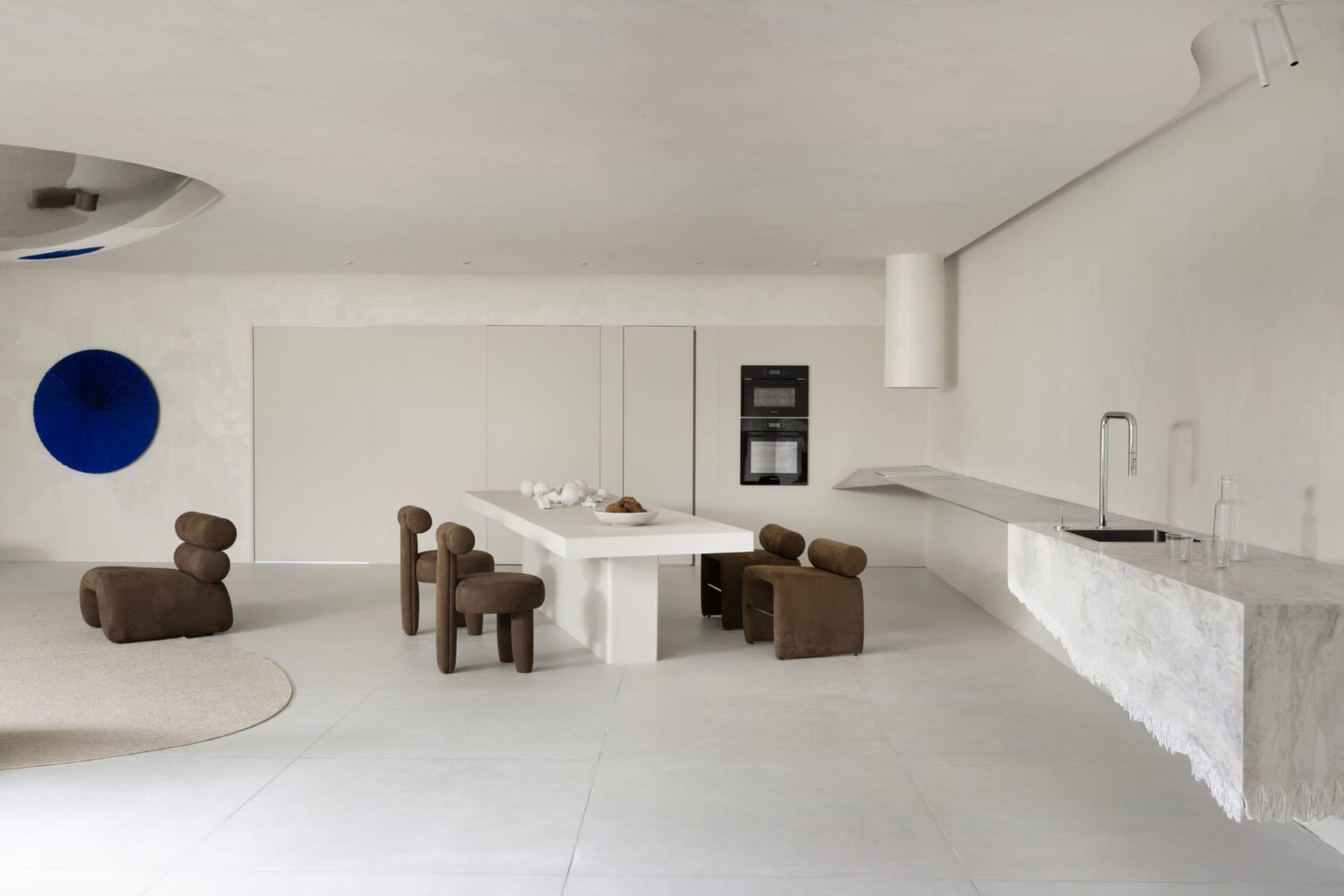A minimalist residential kitchen with a concrete matte white table and chairs, sleek white appliances, and a modern blue roun