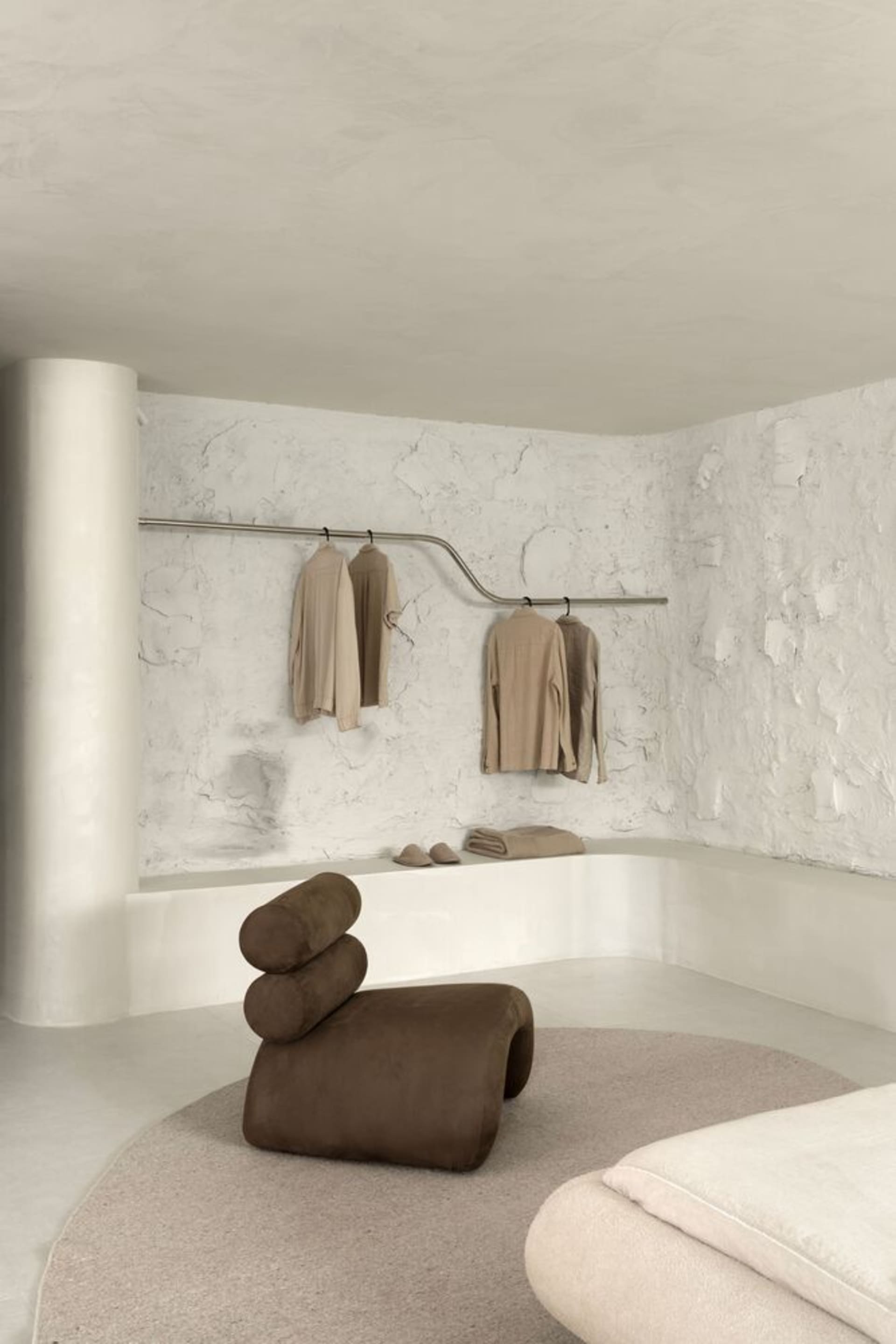 Modern interior with a white wall and concrete texture, featuring a clothing rack with beige garments and a brown sculptural 
