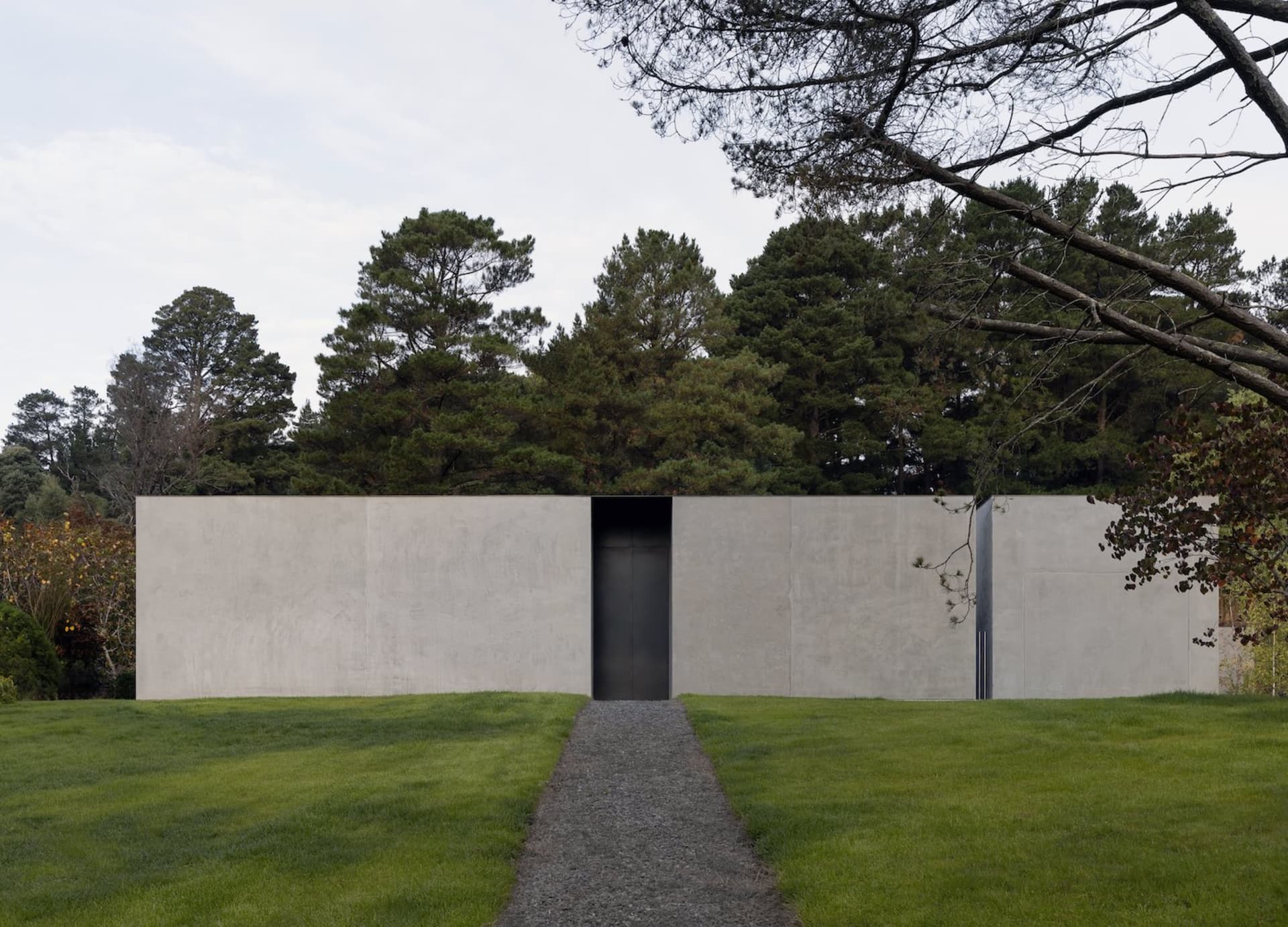 A modern, minimalist building exterior made of gray concrete with a single dark doorway surrounded by green grass and dense t