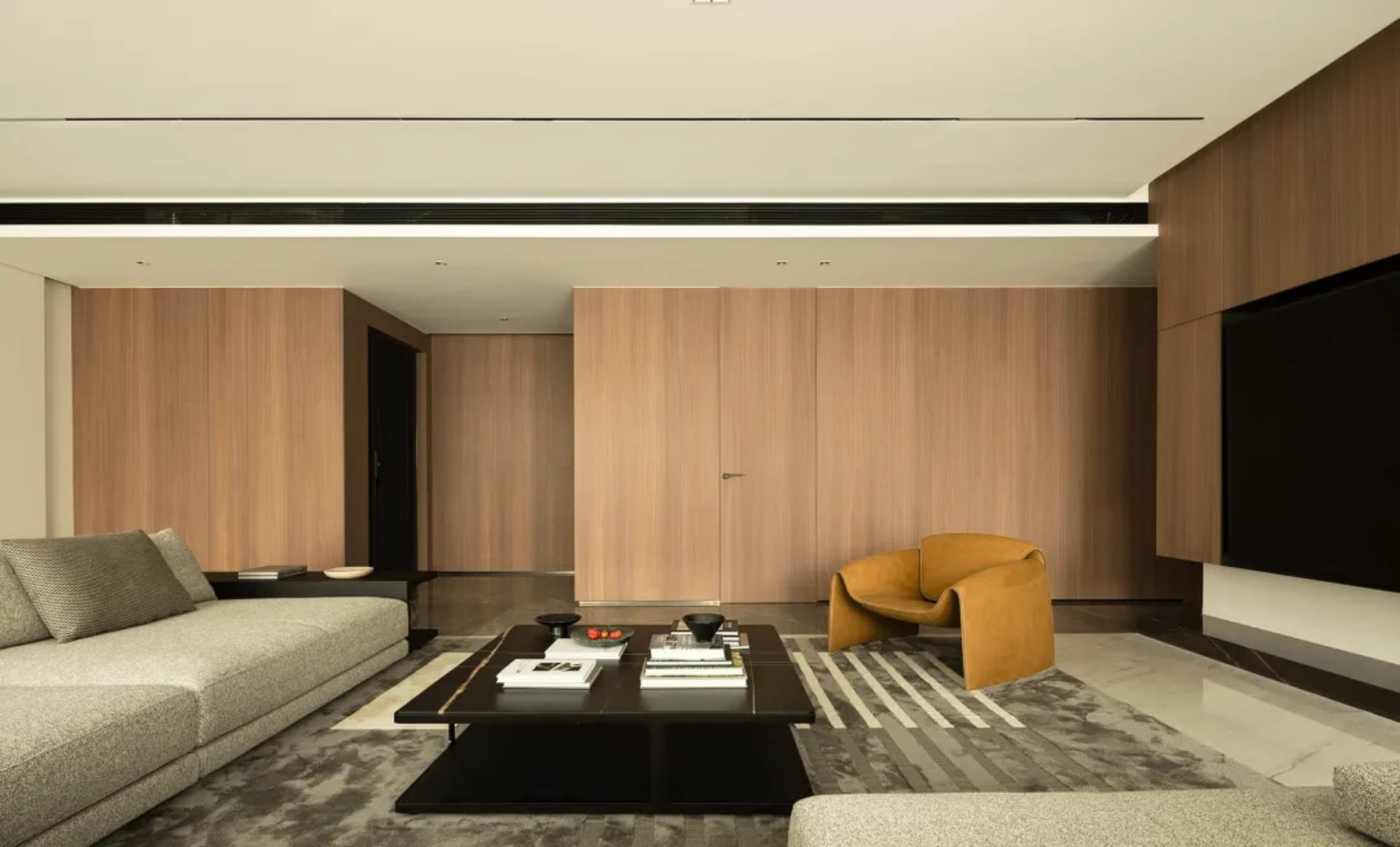 Modern living room with a wooden wall, a beige sectional sofa, an orange armchair, a coffee table with books, and a large scr