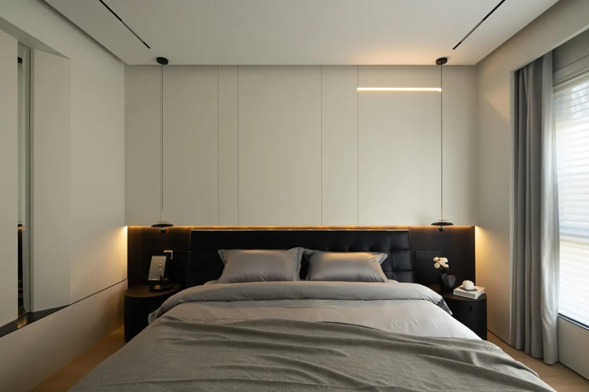 A modern bedroom with minimalist design, featuring a bed with grey bedding and black headboard, illuminated by a linear light