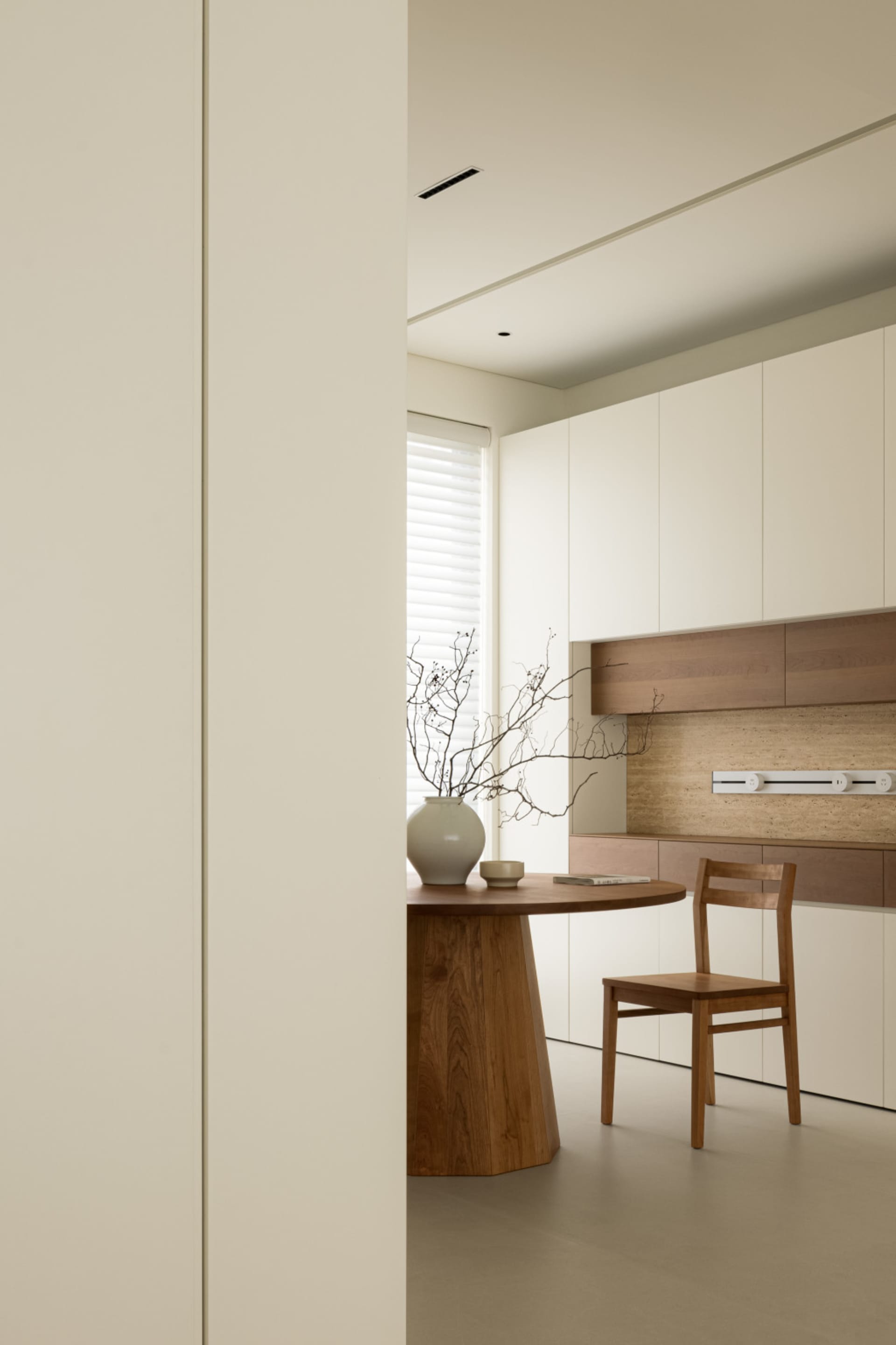 Modern minimalist kitchen with a wooden table and chair, white cabinets, and a vase with branches on the table.
