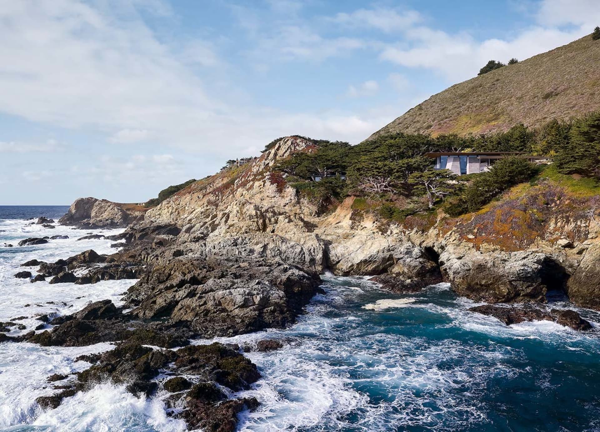 A modern house built into a rocky cliff surrounded by the ocean with greenery.