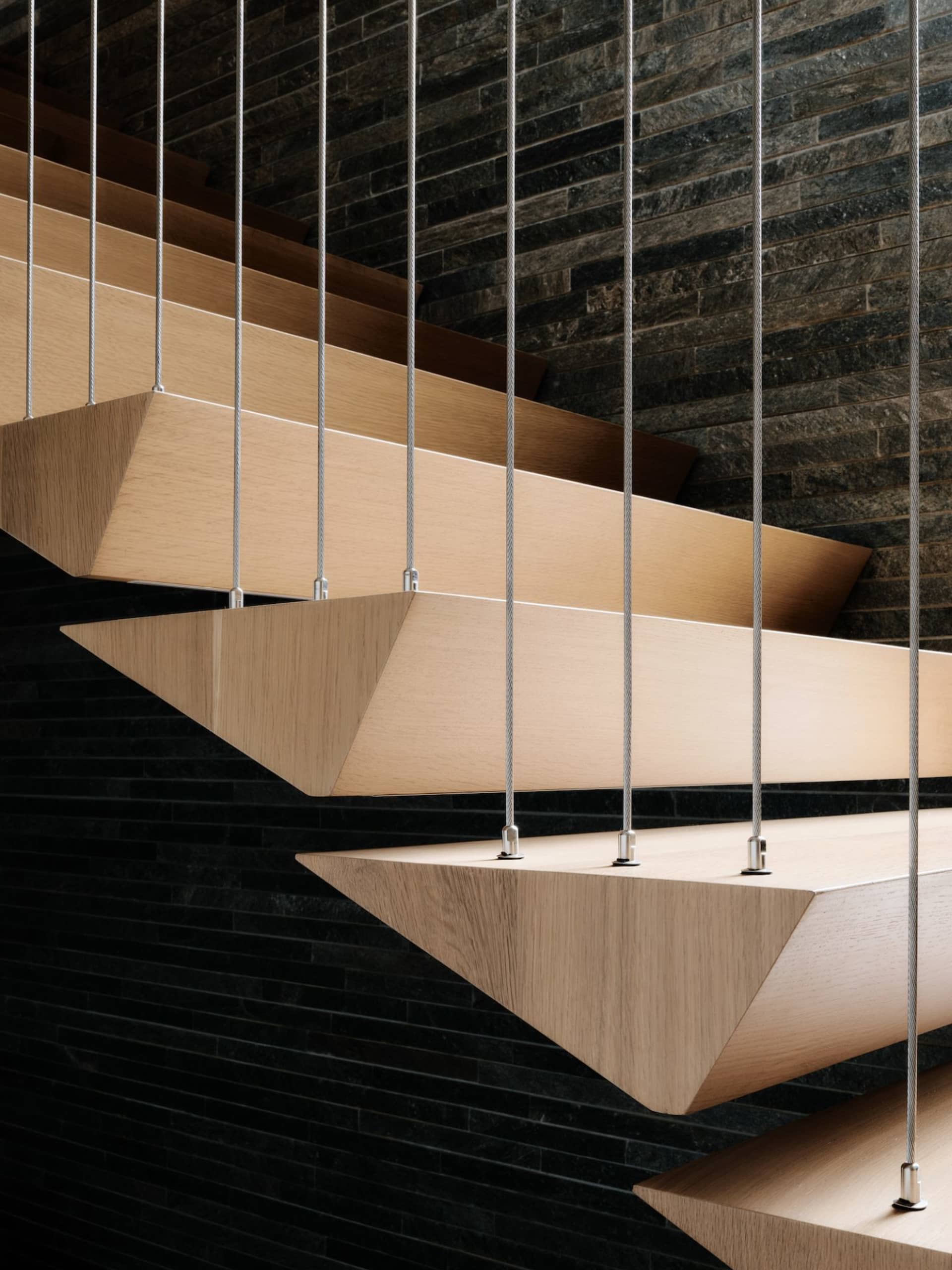 Modern floating staircase with wooden steps hanging by metal cables against a dark brick wall