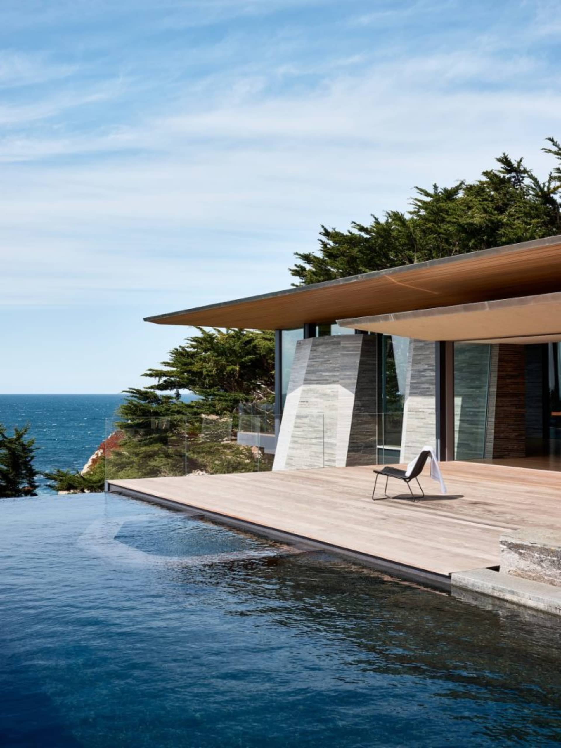An infinity pool with a modern home exterior featuring glass walls and a wooden deck overlooking the sea.