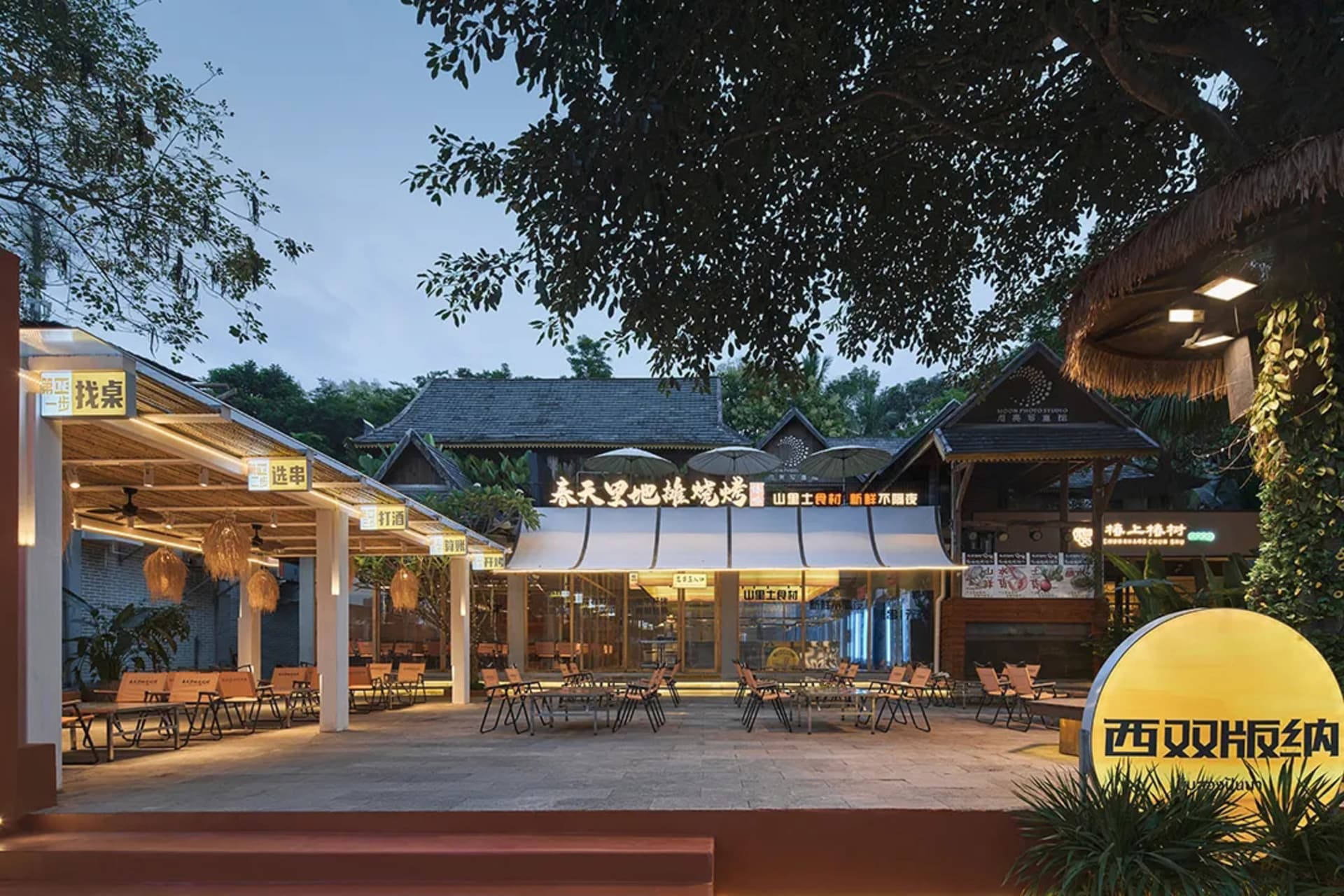 An outdoor seating area of a restaurant with concrete pavement, wooden tables, and glass umbrellas.