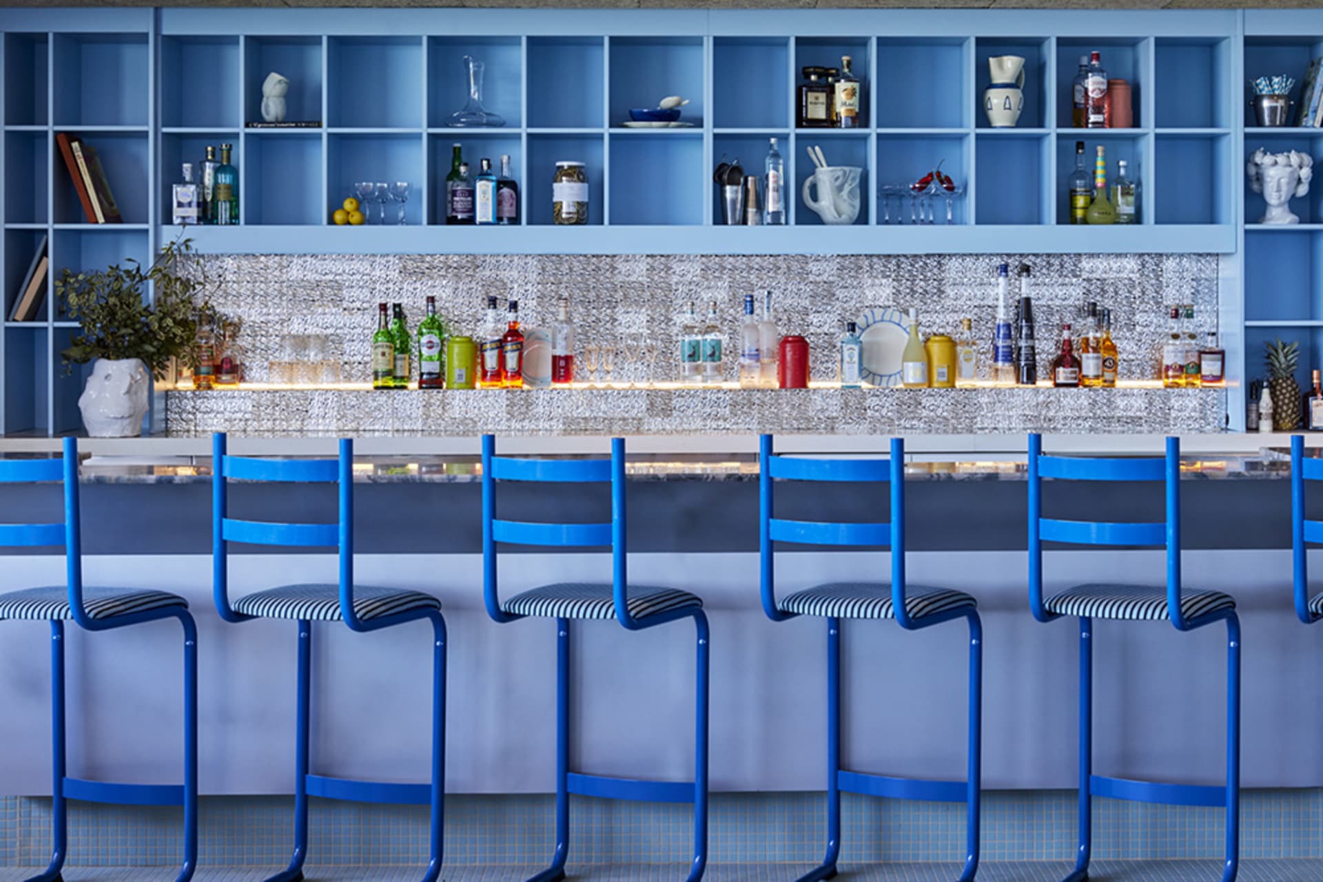 Blue chairs and metallic bar with shelves of bottles and glasses in a bar interior setting