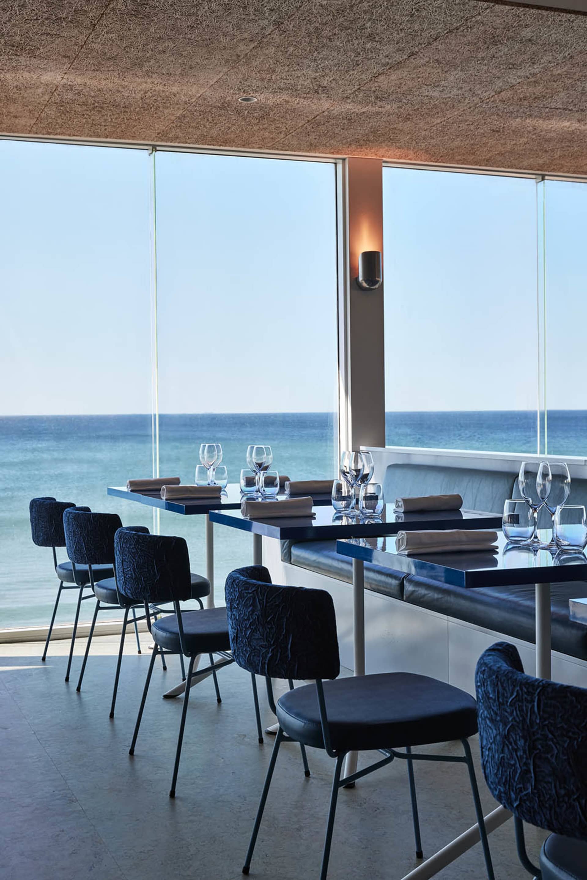 A modern restaurant interior featuring tables, chairs, and wine glasses with a glass wall overlooking the ocean.
