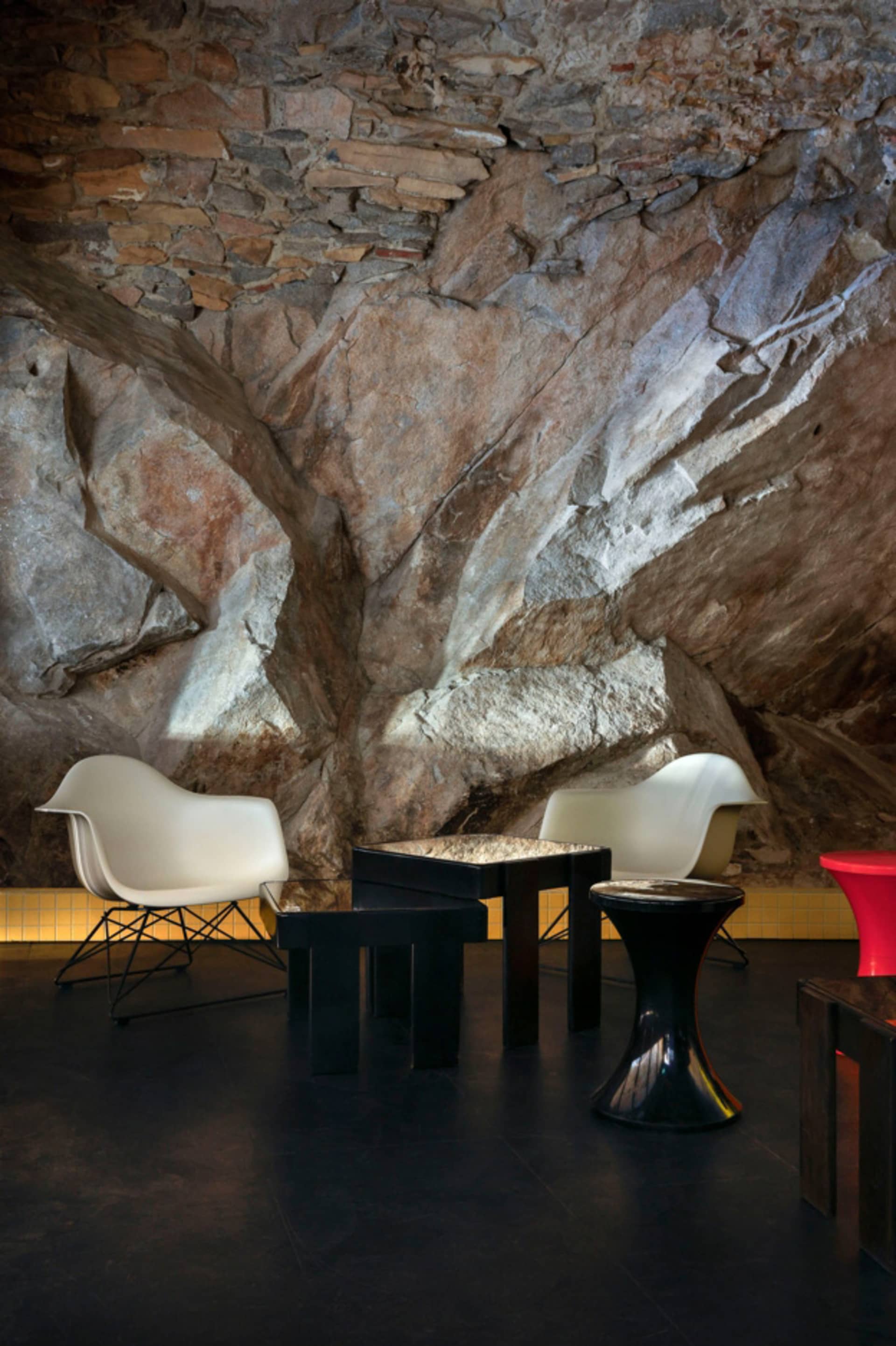 A modern industrial living room with rock walls, white chairs, and black tables and stools.