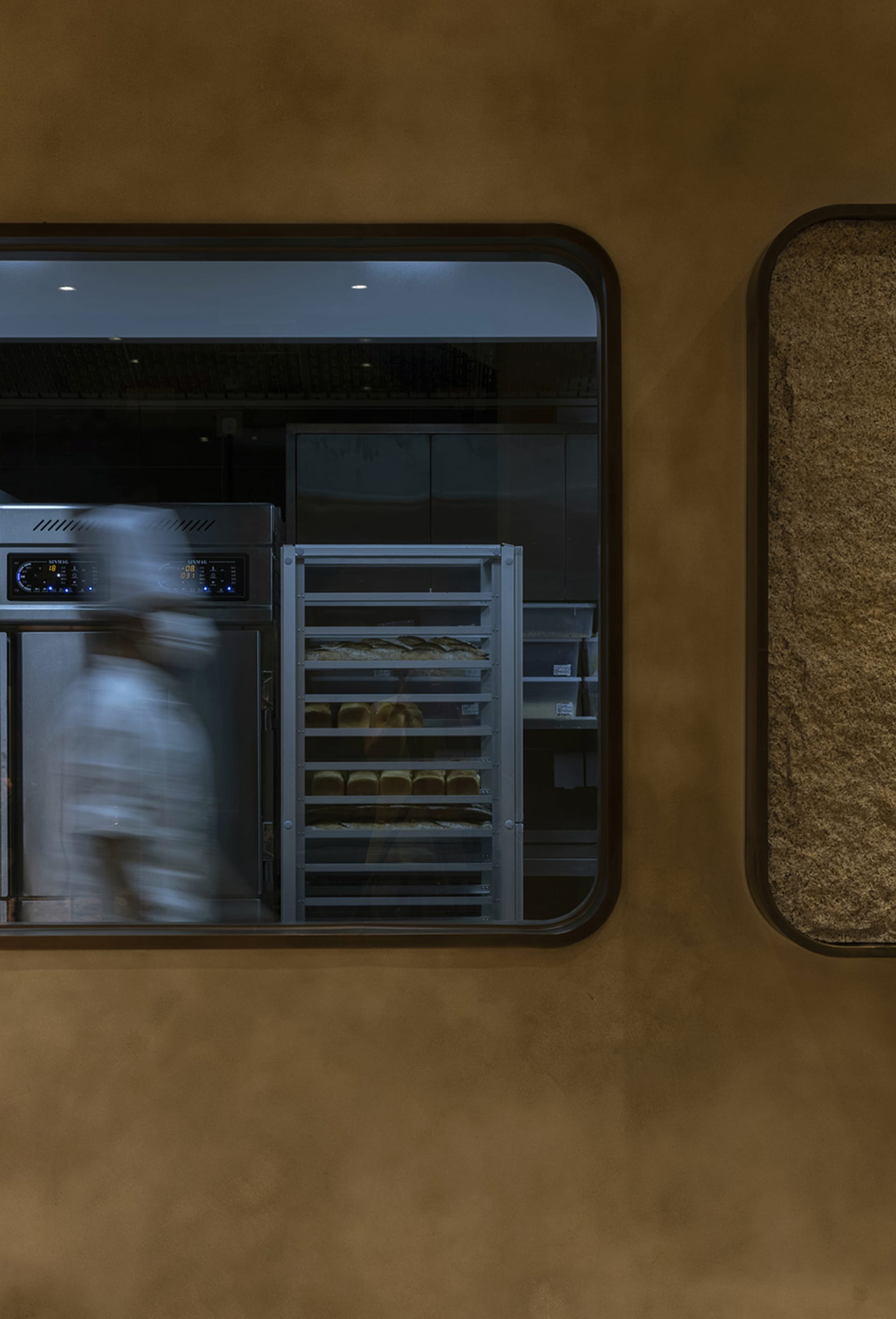 A modern restaurant kitchen with metal shelves and a window showing a baker working in the background.