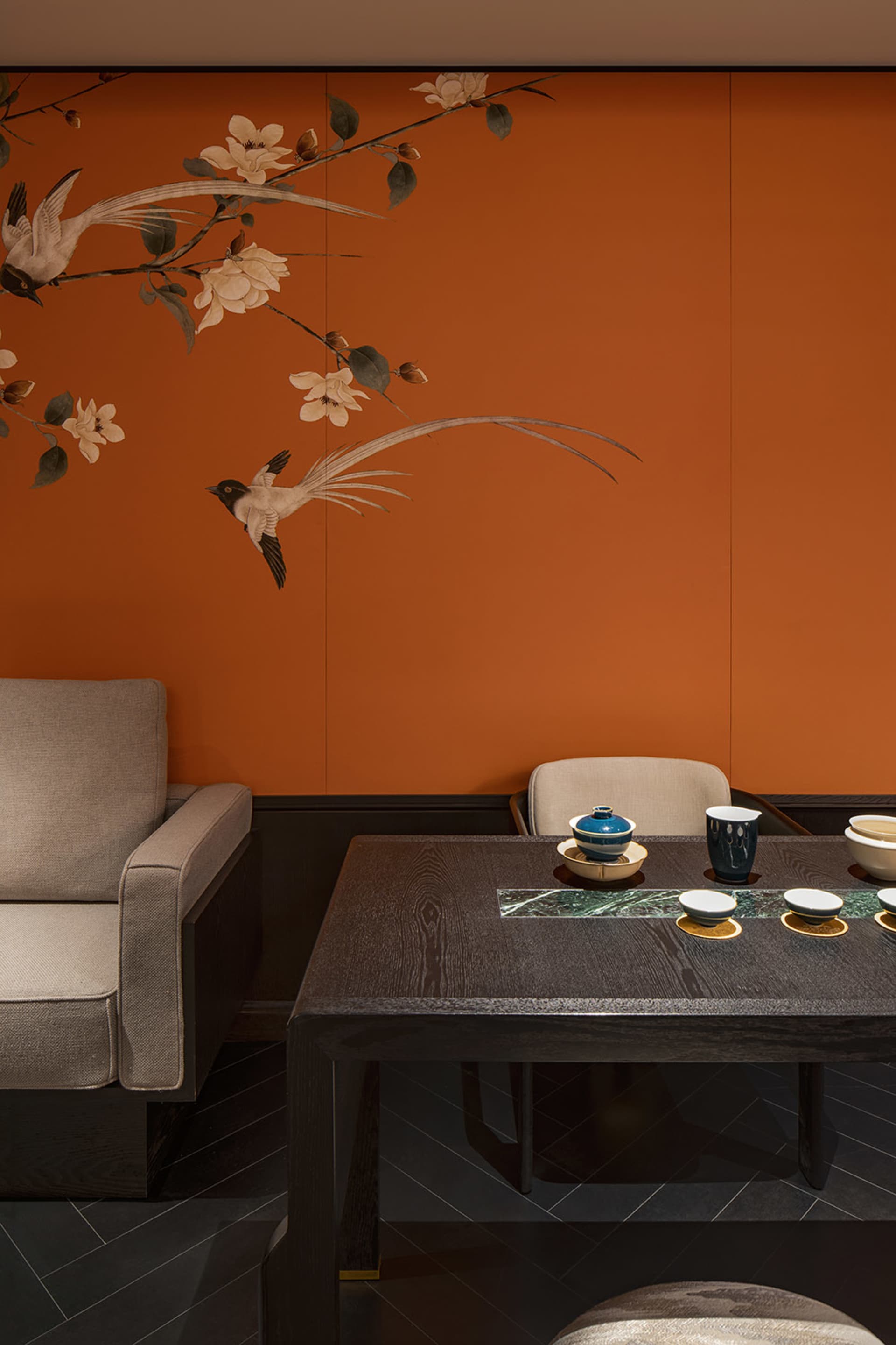 A modern tea room with a dark wooden table and chairs, featuring a dark orange wall with floral and bird decorations and blac