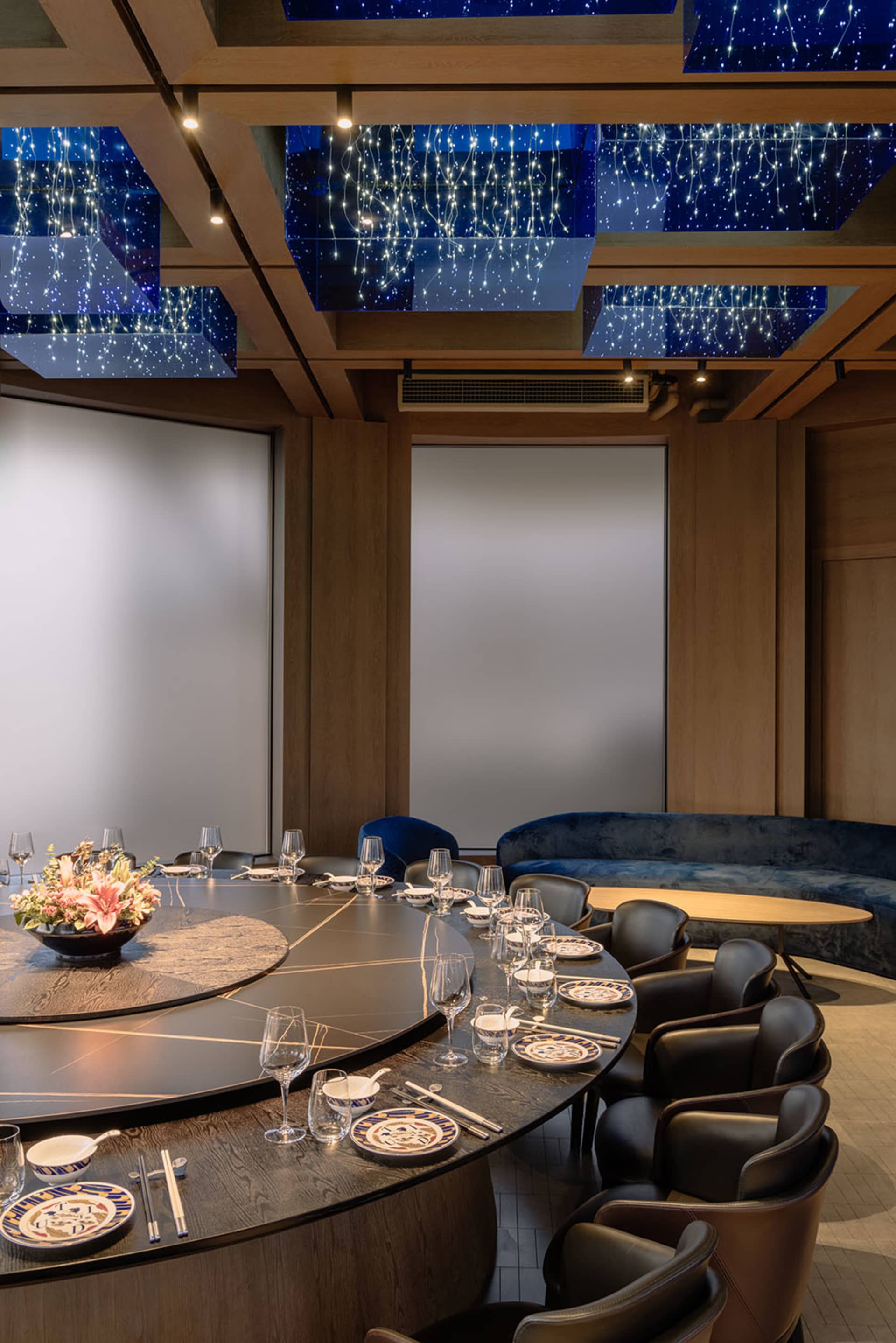 A modern restaurant interior with a wooden round table and chairs, intricate ceiling lights, and a central floral centerpiece